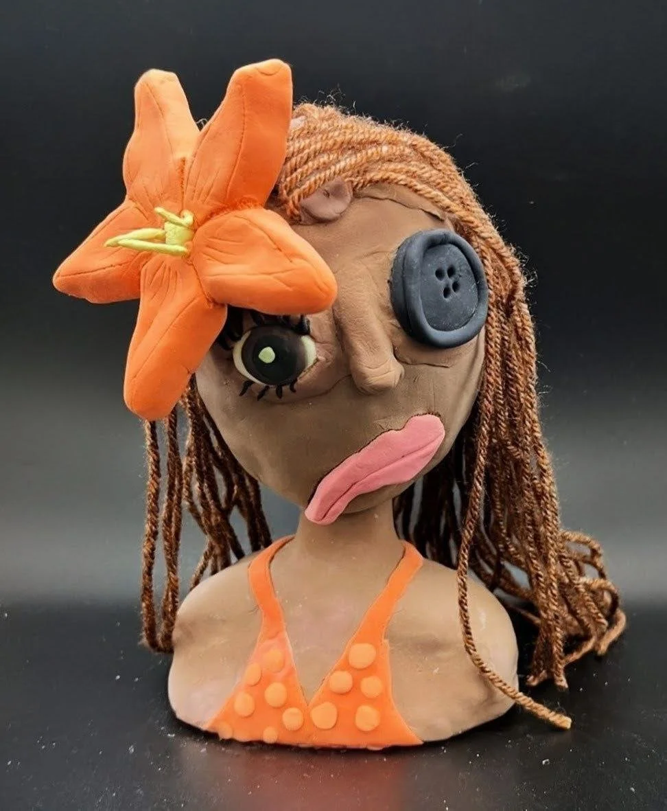 A handmade sculpture of a woman with brown skin, long brown hair, and large exaggerated facial features, decorated with an orange flower on her head, wearing an orange top, and exhibiting a playful, whimsical expression.