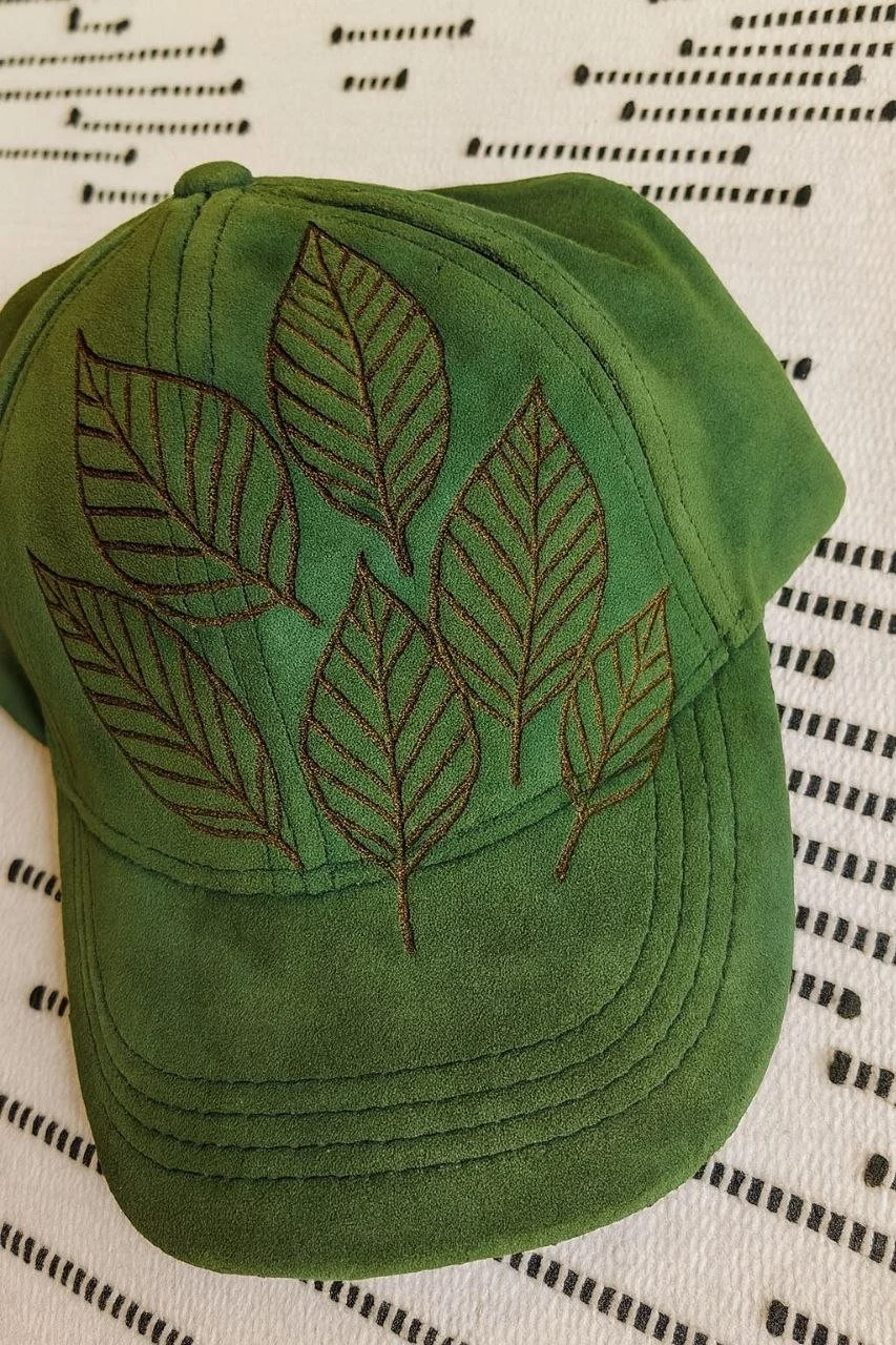 Green baseball cap with embroidered outline of leaves on the front, placed on a white fabric with black dashed linear patterns.