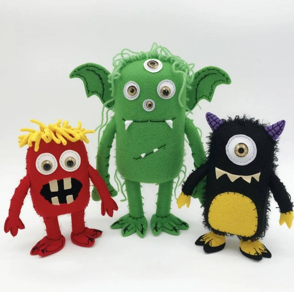 Three plush monster toys with googly eyes against a white background. The largest green monster has three eyes, two bat wings, and white fangs. The red monster has yellow yarn hair, two eyes, and a black mouth with white teeth. The black monster has purple horns, one eye, and a yellow belly.