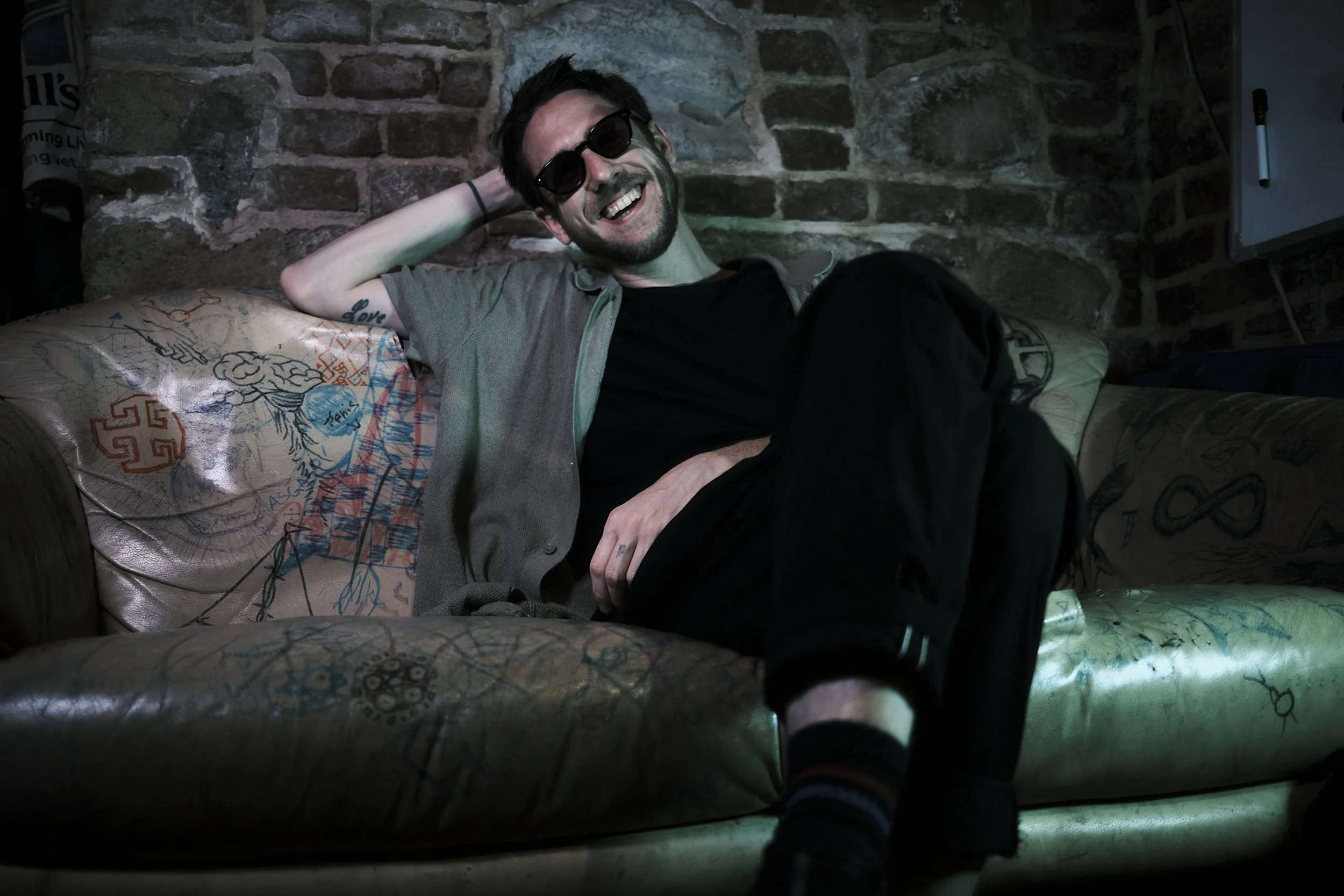 Junger Mann mit dunklen Haaren, Brille und grauem Hemd sitzt lächelnd auf einem bunten, graffiti-verzierten Sofa vor einer Backsteinwand, in einem dunklen Raum.
