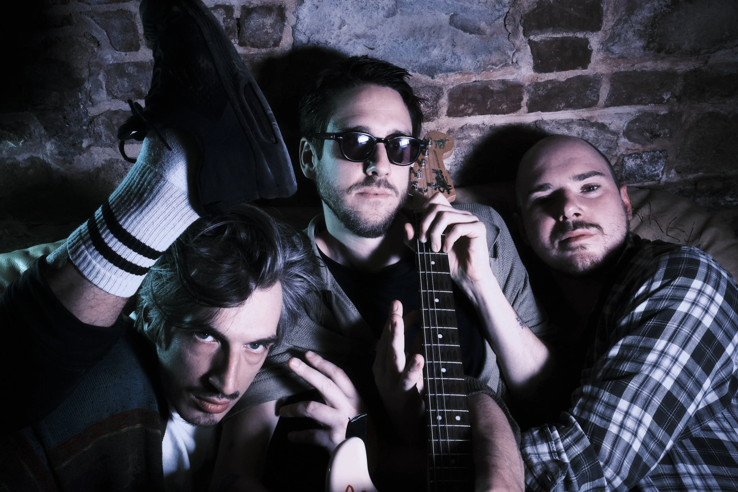 Drei junge Männer posieren in einer gemütlichen, steinernen Umgebung; einer hält eine E-Gitarre, der andere trägt Sonnenbrille, alle zeigen einen entspannten, rebellischen Stil.