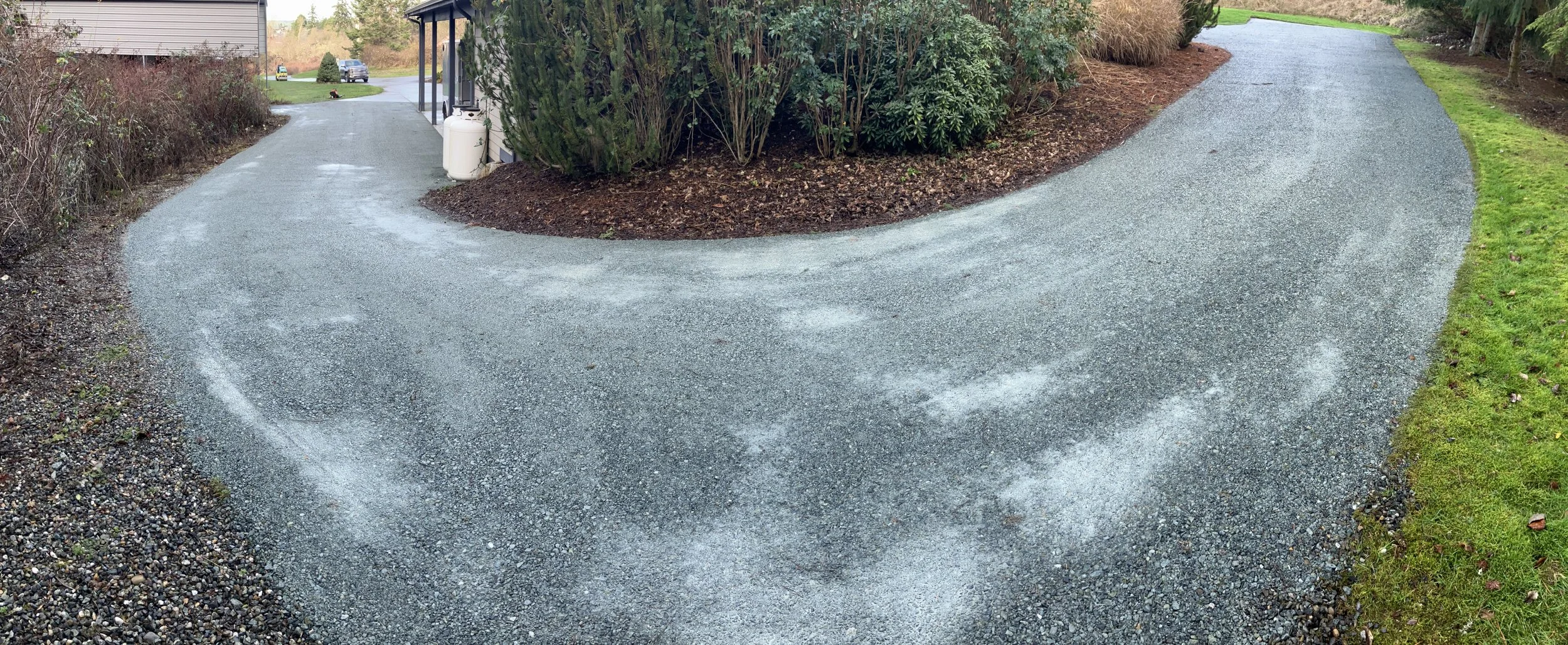 A curved gravel driveway with a small patch of gravel on the left side and a grassy lawn on the right, surrounded by bushes and trees.