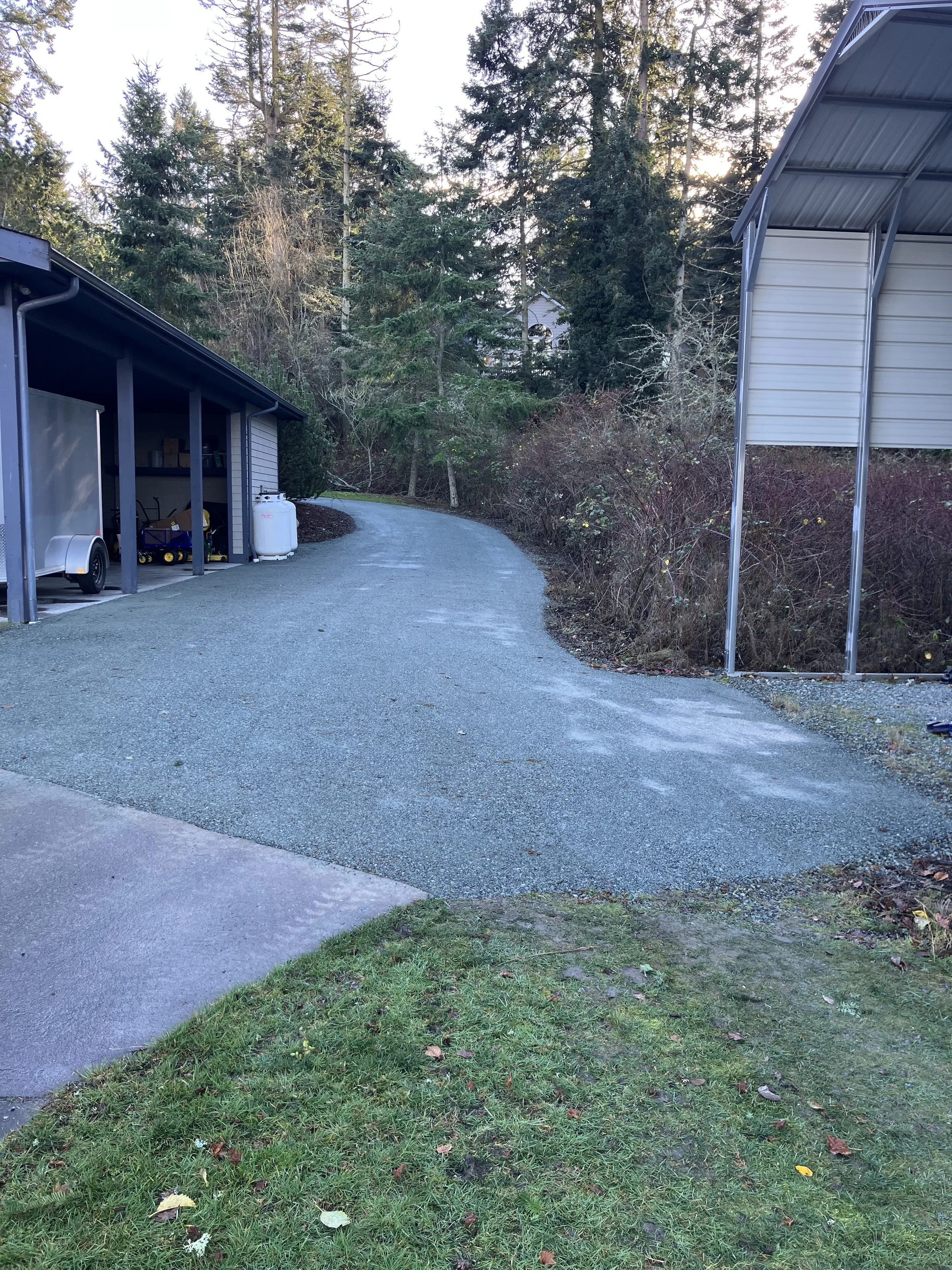 A resurfaced gravel driveway curves between two metal structures, with trees and bushes in the background and a grassy area in the foreground.