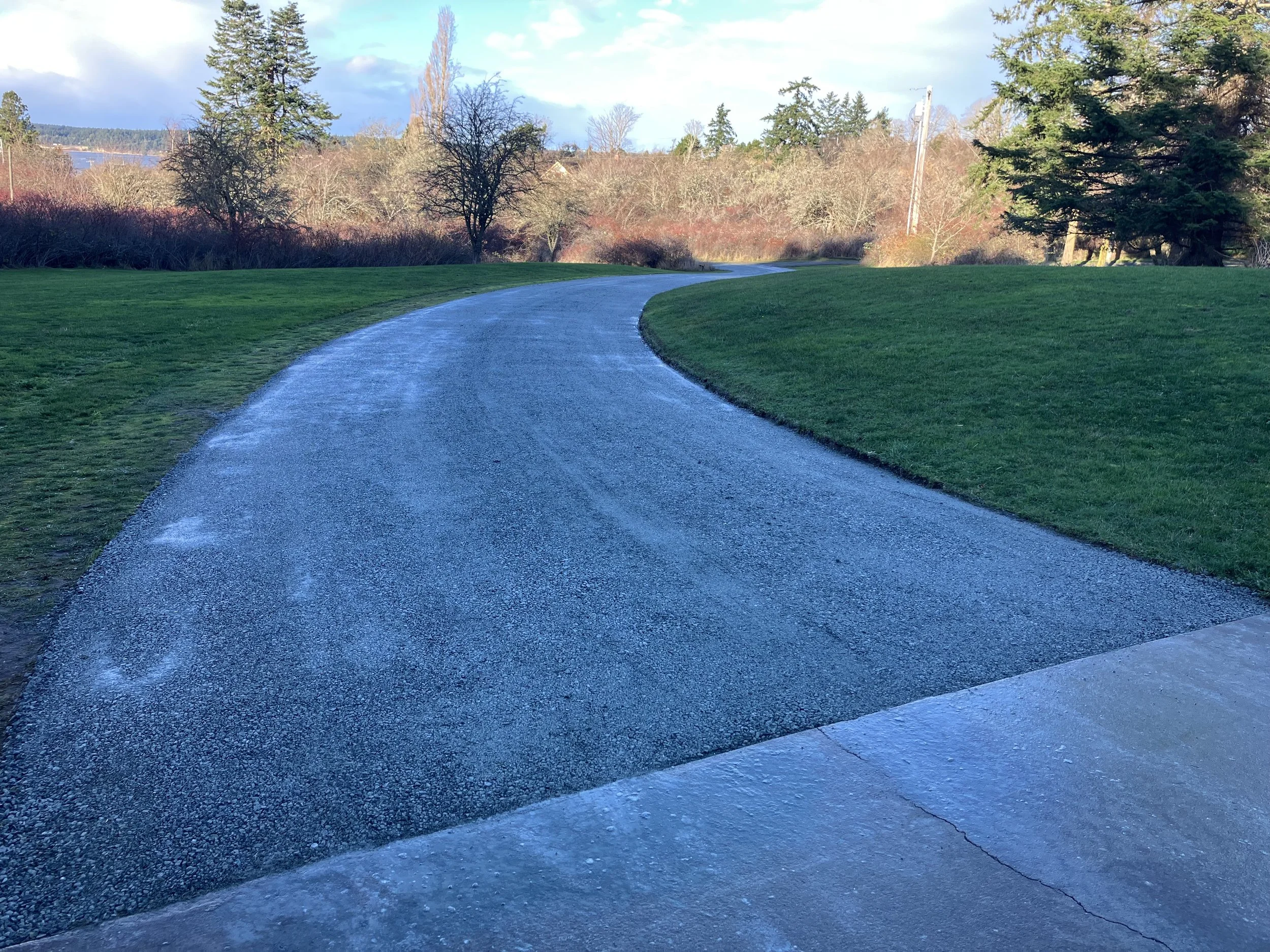 A winding winding new gravel driveway with a grassy lawn on both sides, leading into a landscape with trees and a partly cloudy sky.