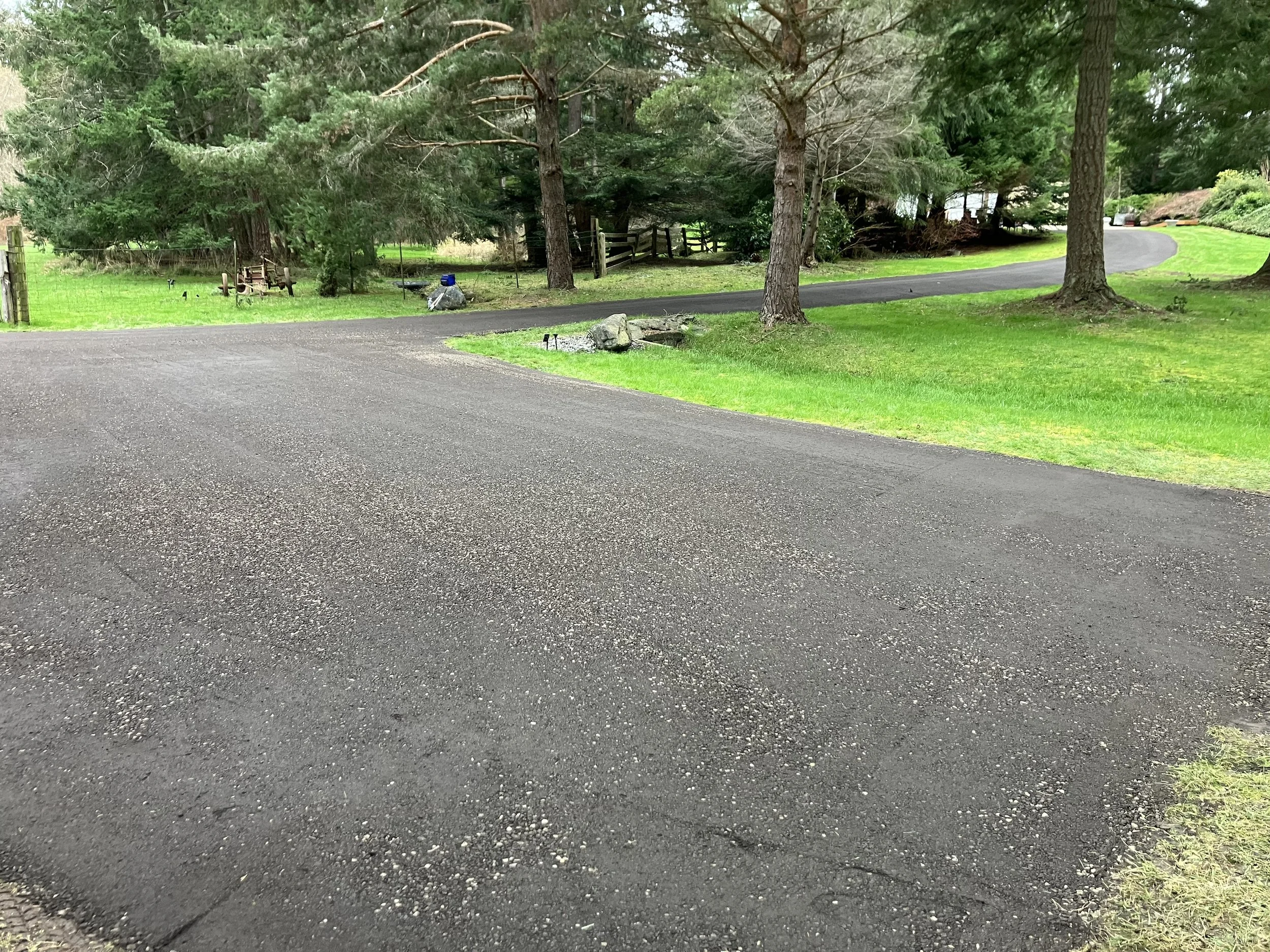 A new resurfaced crushed recycled asphalt gravel driveway curves through a green yard with grass and trees in a residential area.