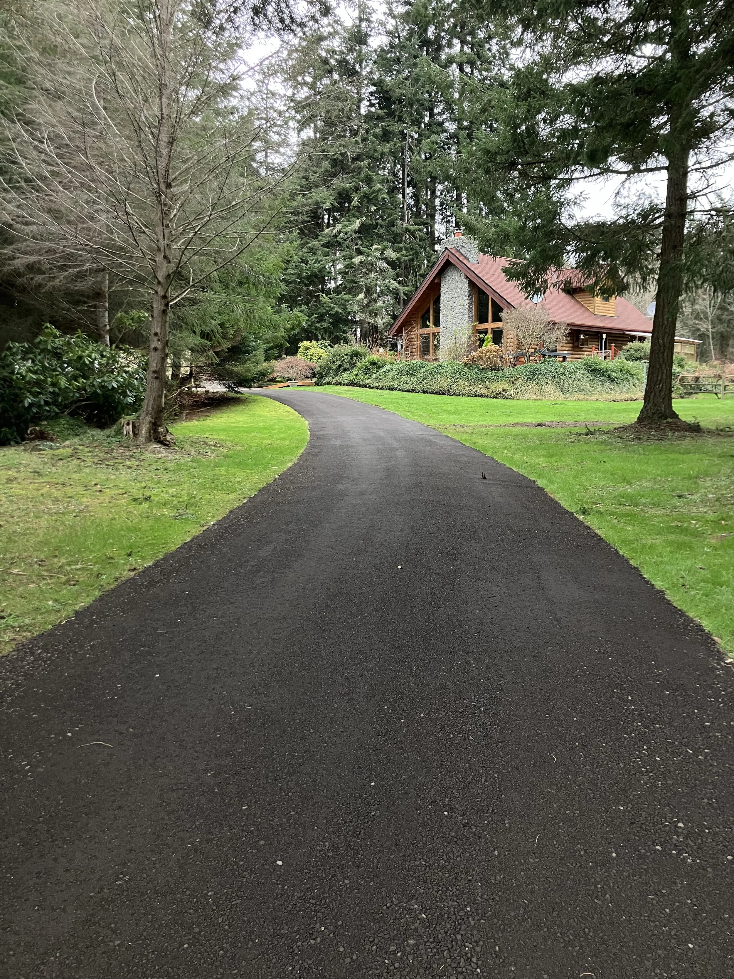 A new resurfaced crushed recycled asphalt gravel driveway winding through a lush green yard towards a log cabin style house with a stone chimney, surrounded by tall trees and landscaped bushes.