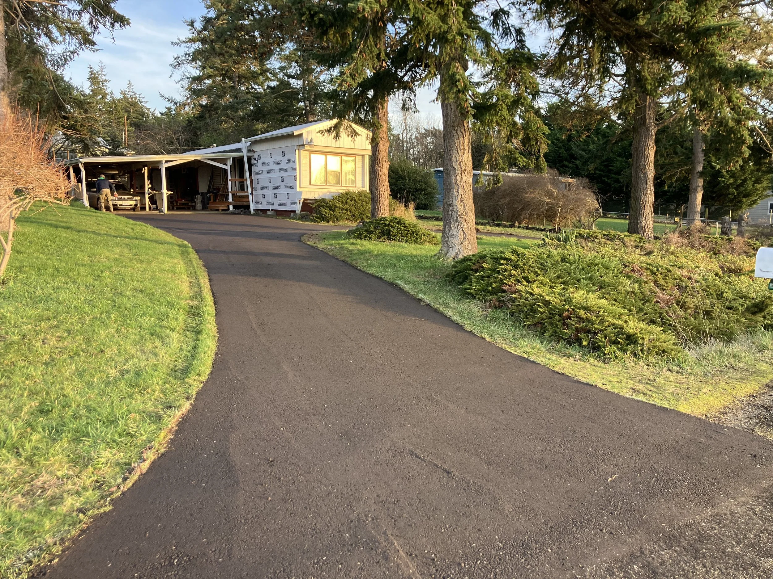 A new refreshed resurfaced crushed recycled asphalt gravel driveway leads to a partially constructed house with a carport, surrounded by green grass, trees, and shrubbery, in a residential area.