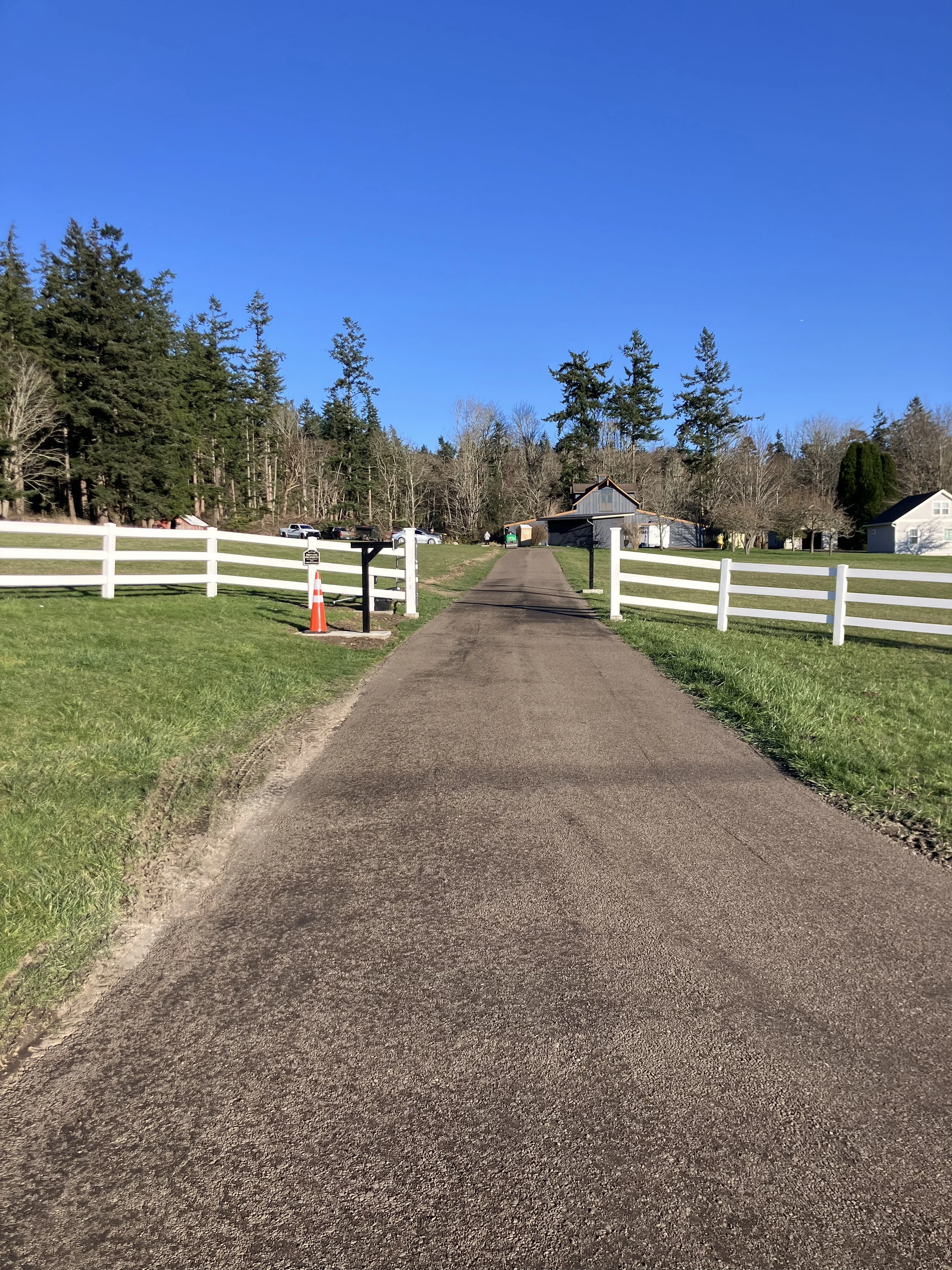 A new resurfaced crushed recycled asphalt gravel driveway with white fences on both sides leading to a house in a rural area under a clear blue sky, with trees in the background.