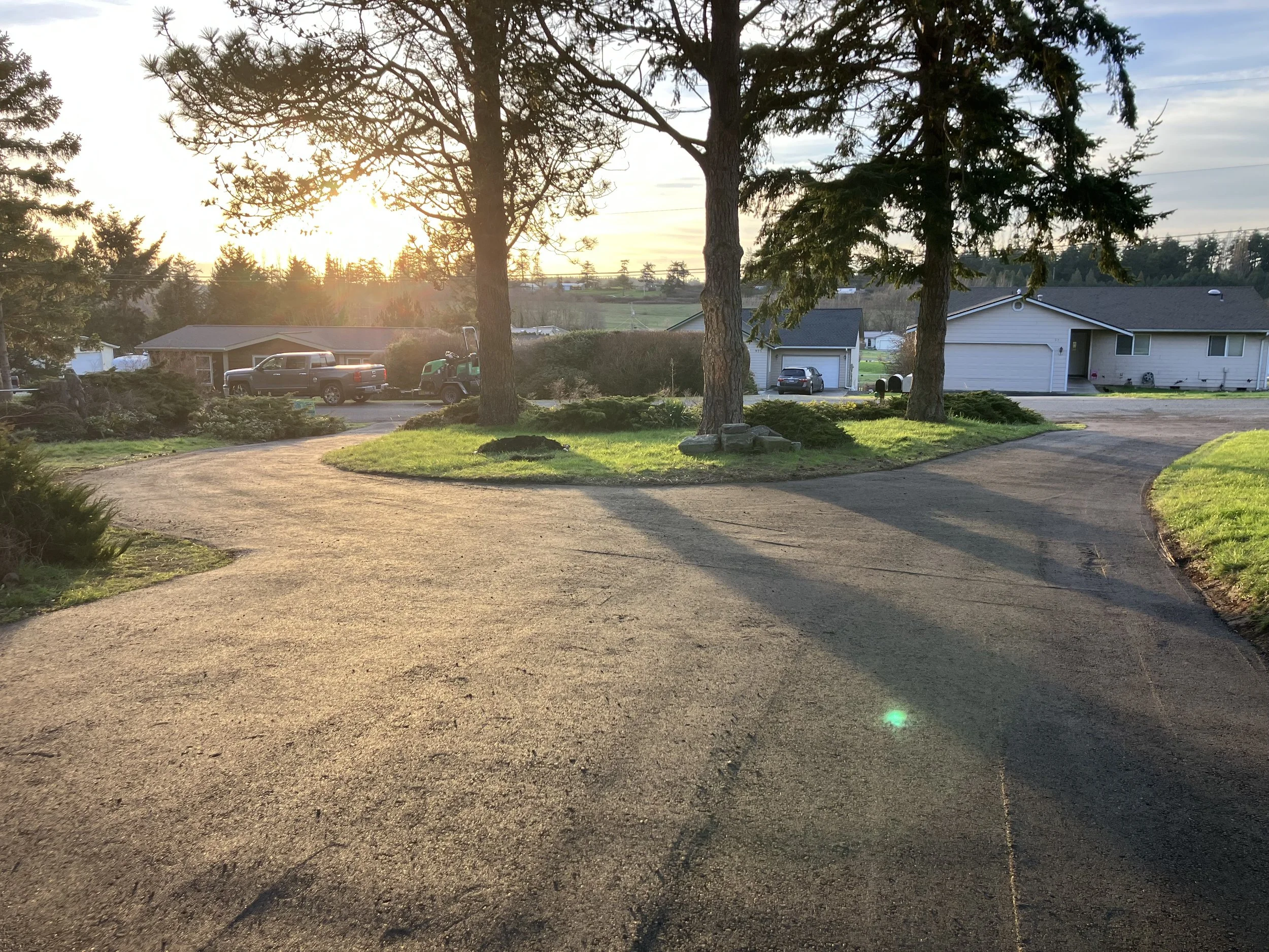 A new resurfaced gravel driveway using crushed recycled asphalt in a residential neighborhood at sunset, with trees, houses, parked cars, and a green lawn.