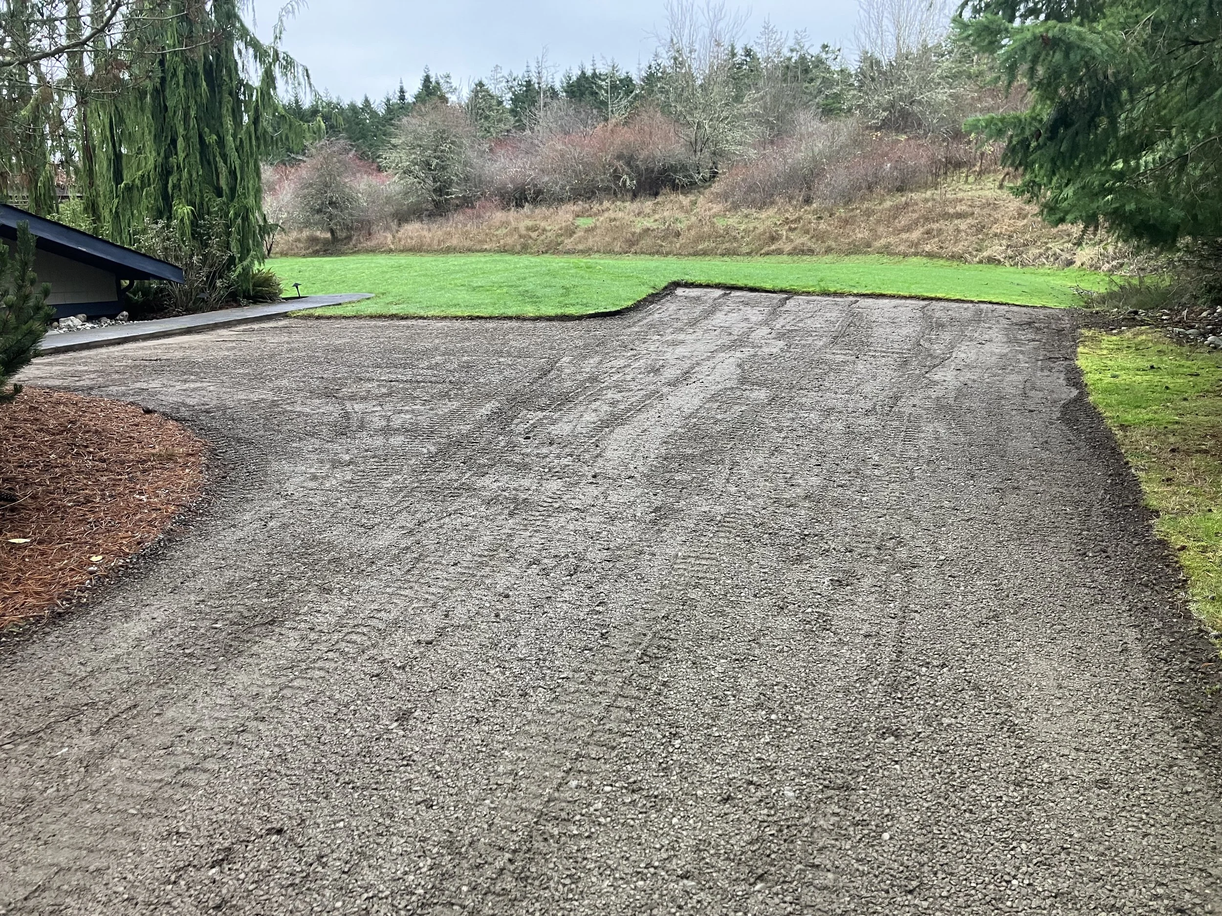 A prepared gravel driveway that has been compacted and is ready for new gravel with green grass and trees on one side and a pine needle flower bed on the other side under a cloudy sky.
