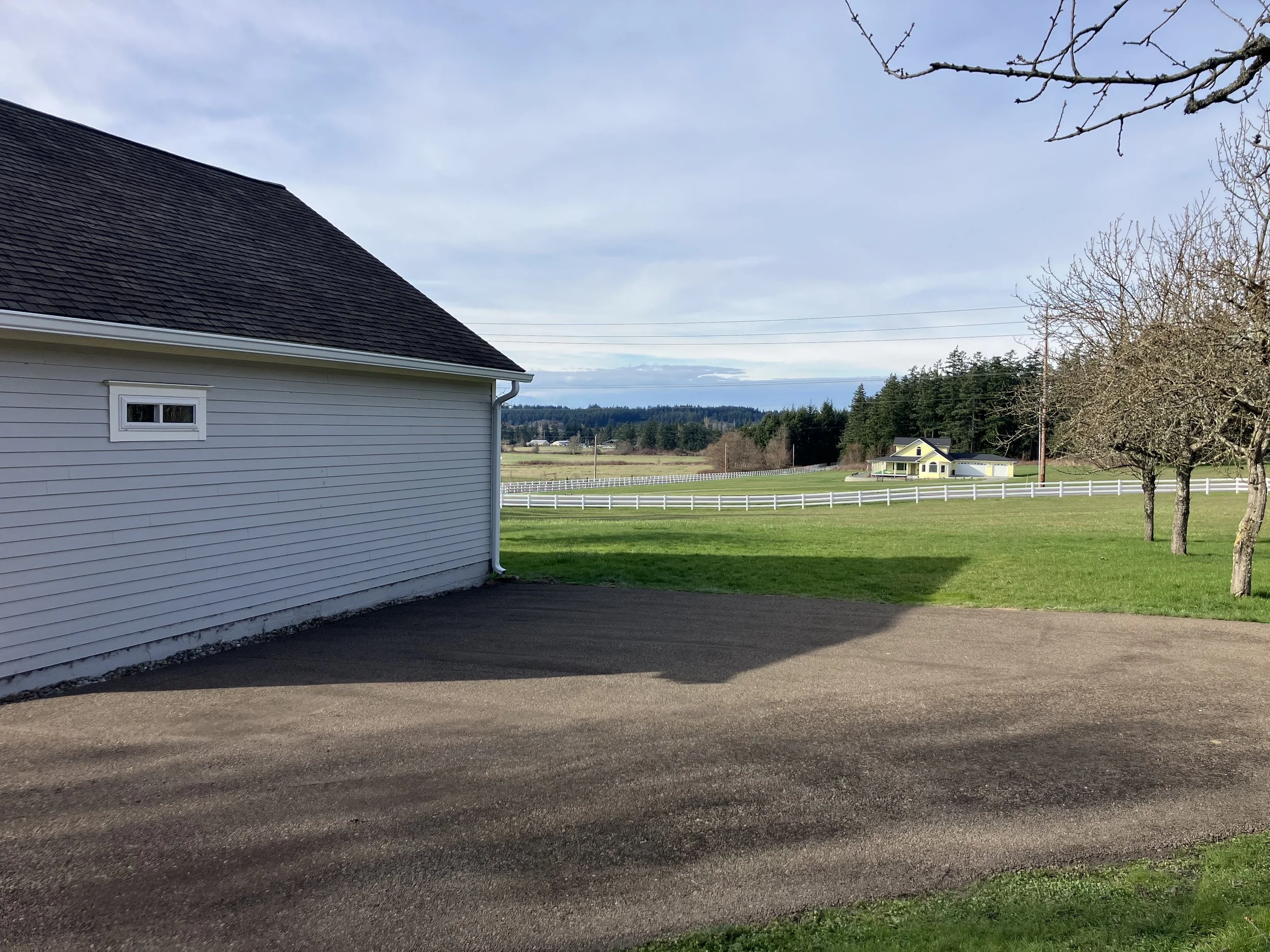 A rural scene with a newly refreshed recycled asphalt gravel driveway and a white house on the left, green grass and trees on the right, and a distant yellow house with white fencing in the background, under a partly cloudy sky.