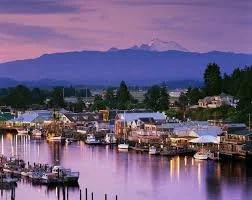 A scenic view of a marina at dusk with boats docked along the water, mountains in the background, and a partly cloudy sky with purple hues.