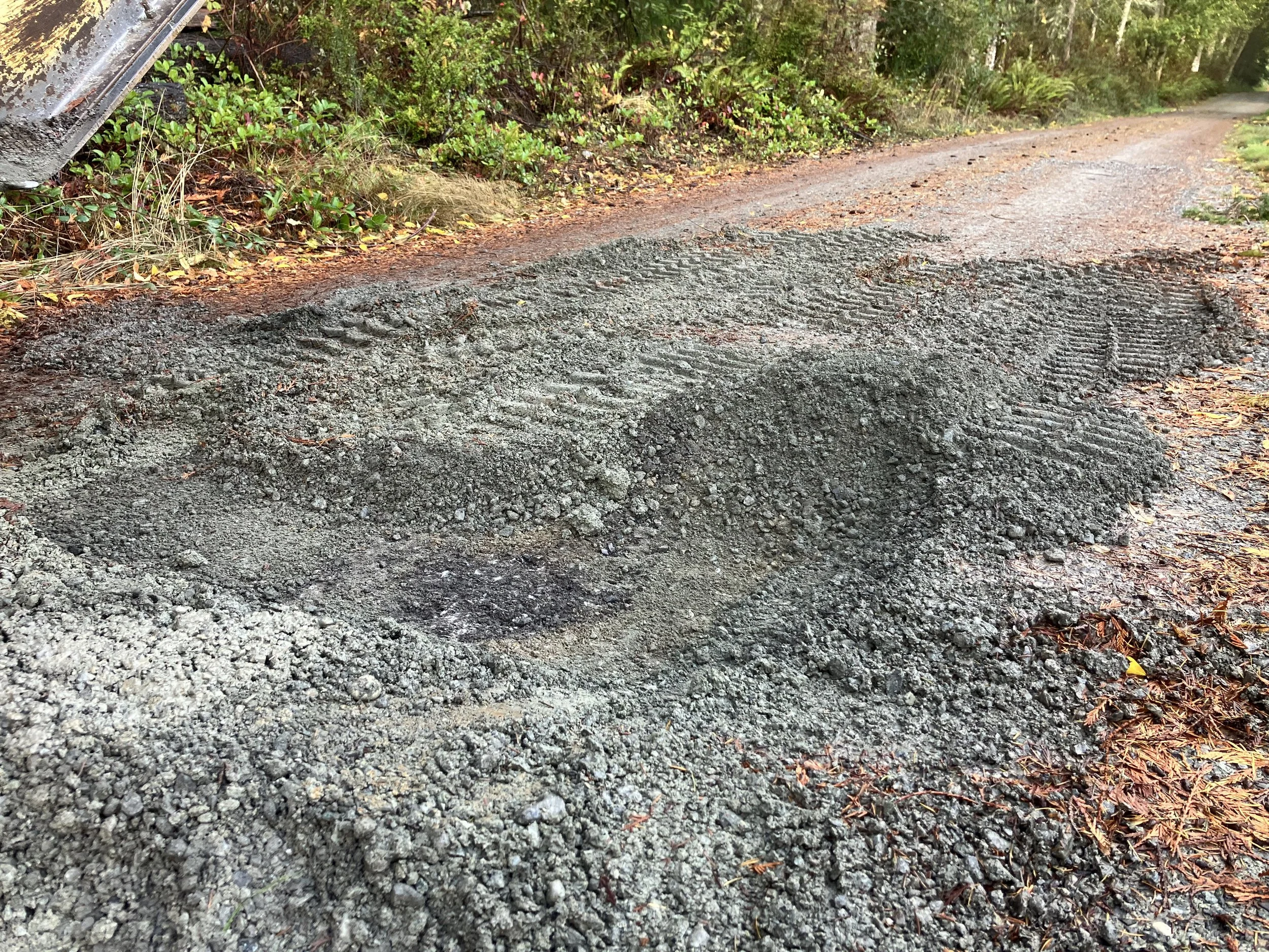 A dirt road with a freshly patched section of gravel, surrounded by green bushes and trees.