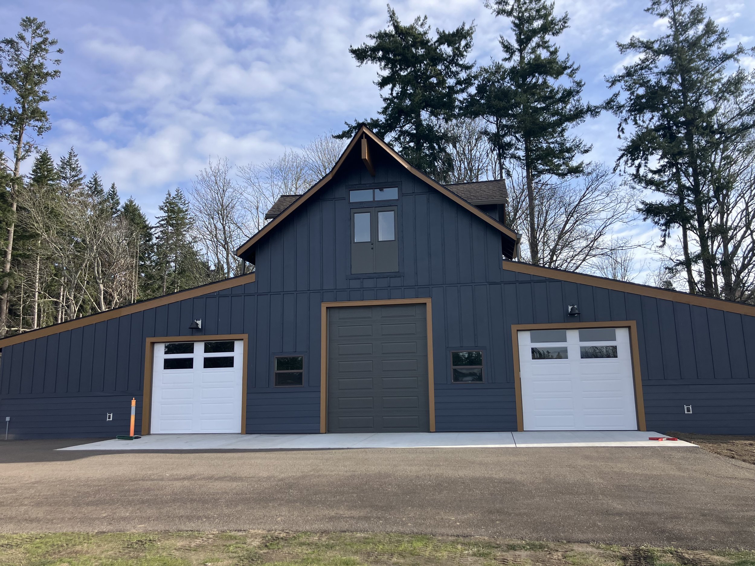 A modern blue barn with three garage doors, surrounded by trees and a partly cloudy sky with a new resurfaced recycled asphalt gravel driveway.