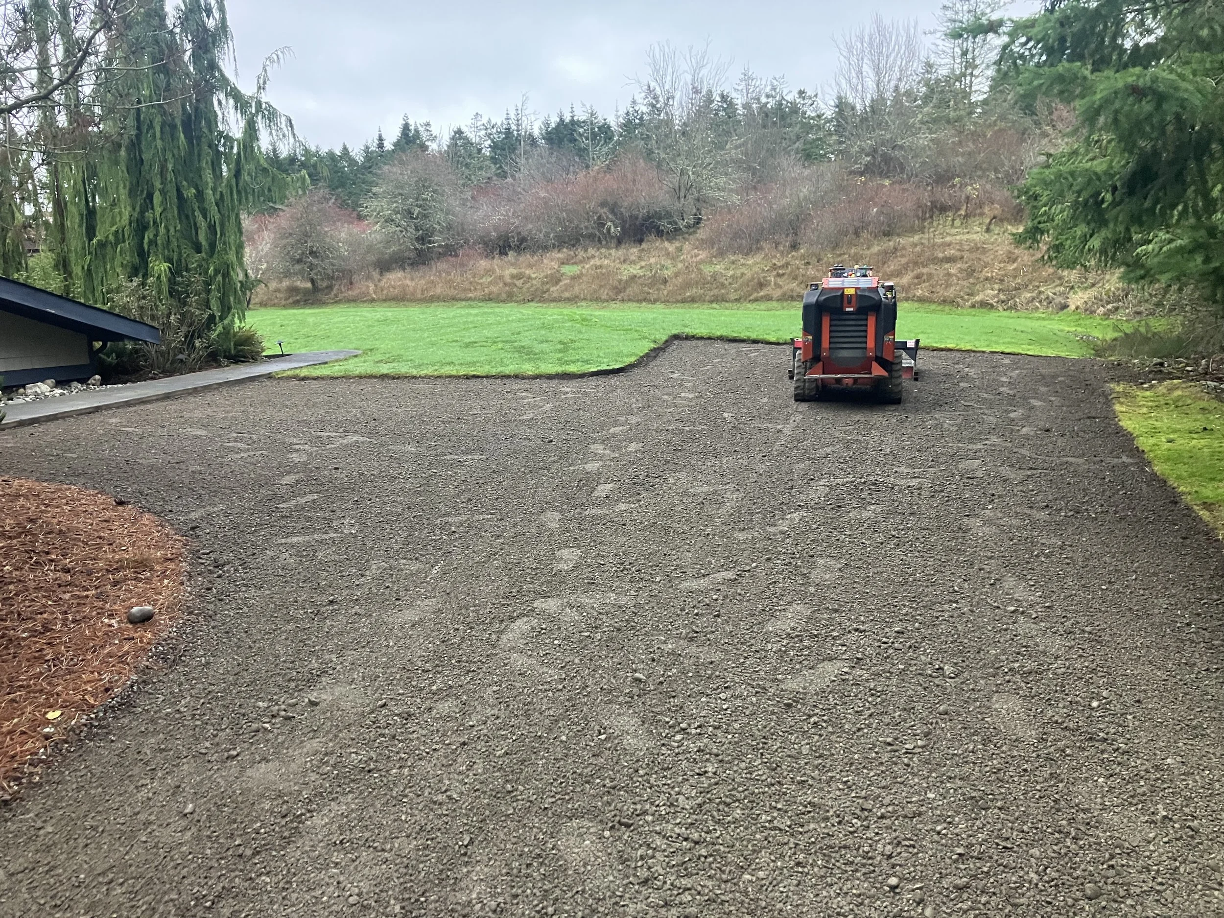 A prepared gravel driveway with green grass on one side and a pine needle bed on the other side under a partly cloudy sky.
