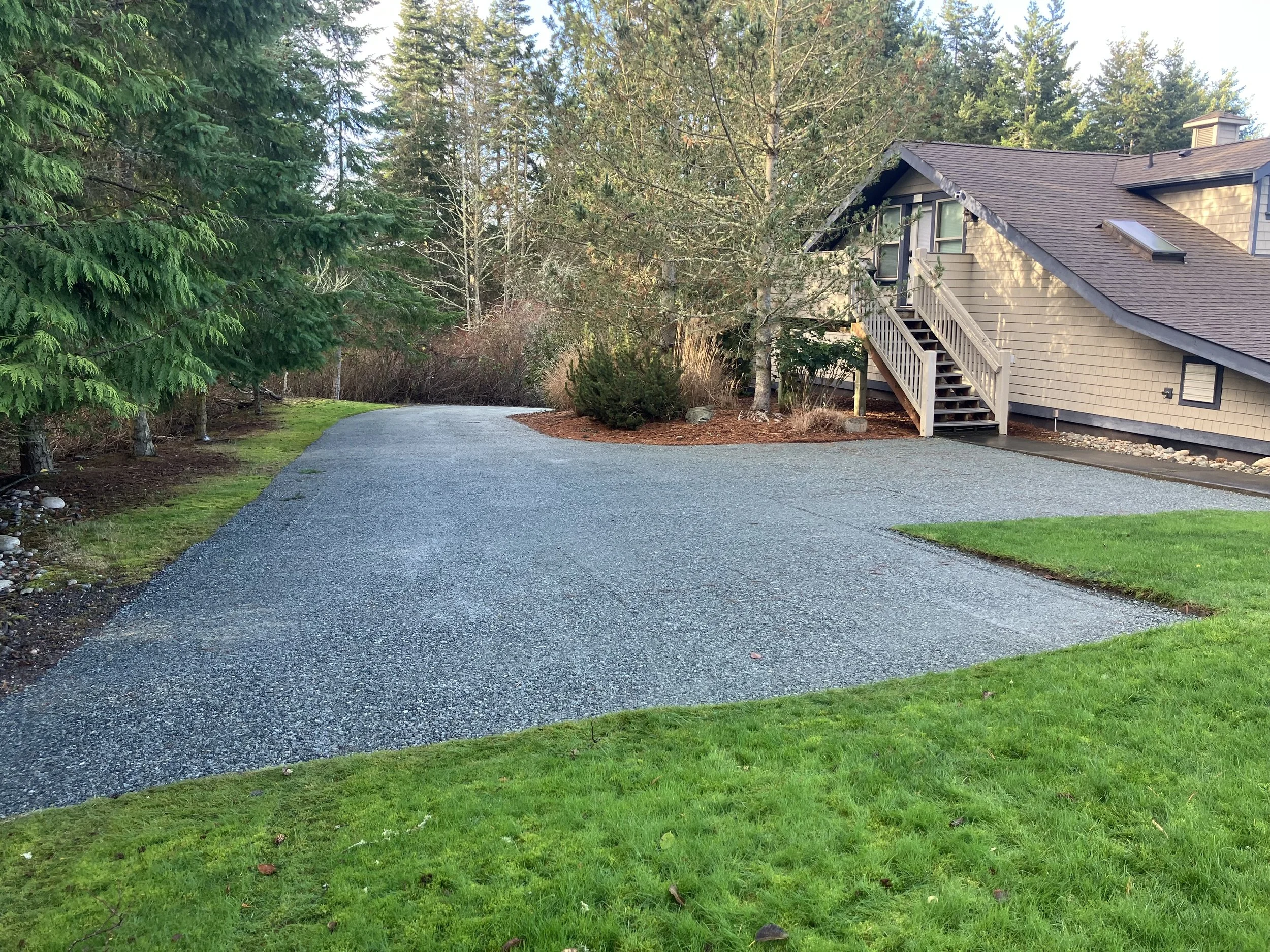 A new gravel driveway curves past a beige house with stairs leading up to a door, bordered by green grass and trees in a suburban setting.