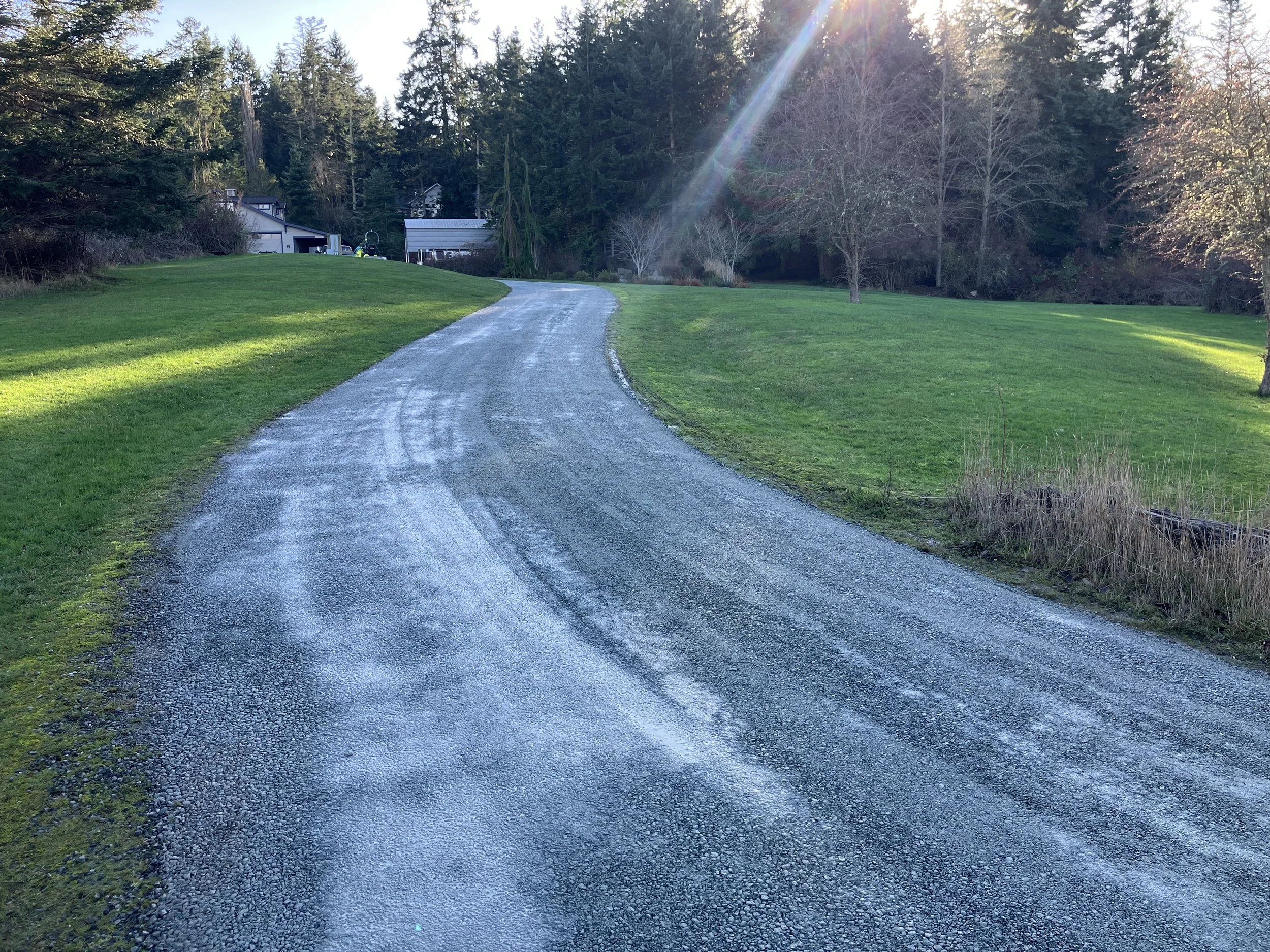 A winding new resurfaced gravel driveway  with no potholes running through a grassy lawn, with trees and houses in the background under a bright sky and sunlight rays.