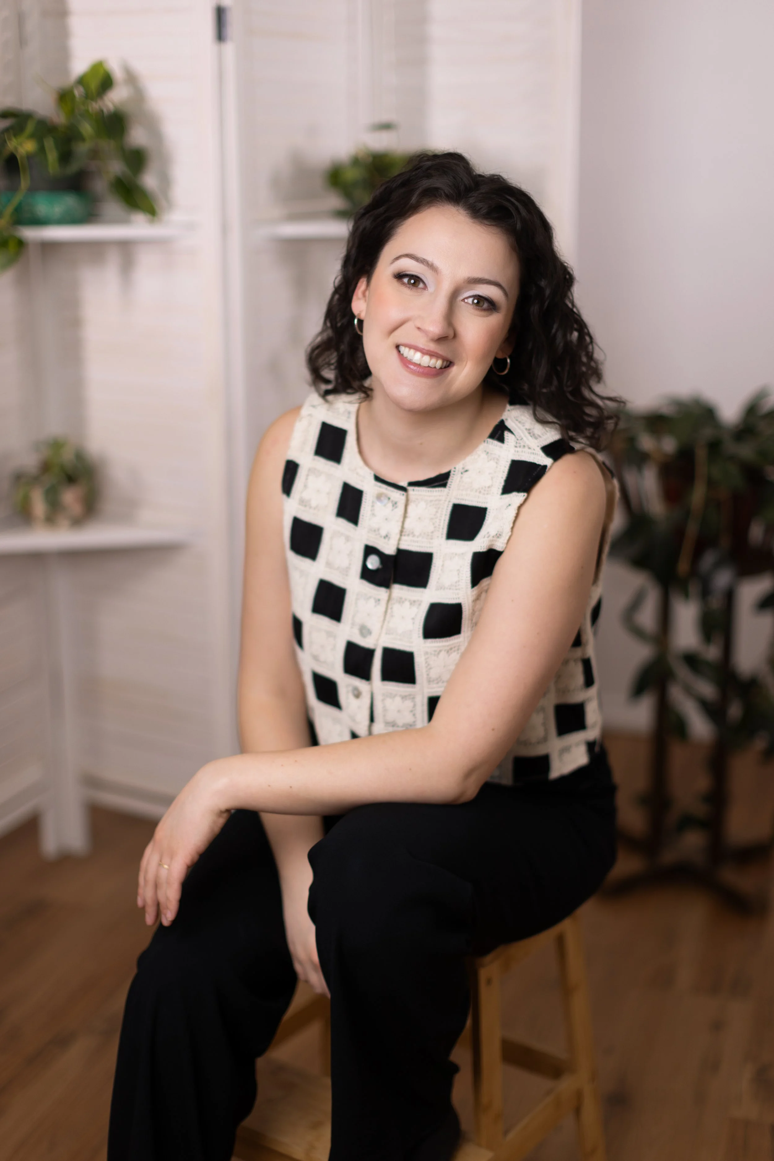 A woman with dark, curly hair smiling at the camera, wearing a black and white checkered sleeveless top, sitting on a wooden chair indoors, with plants and a white background behind her.