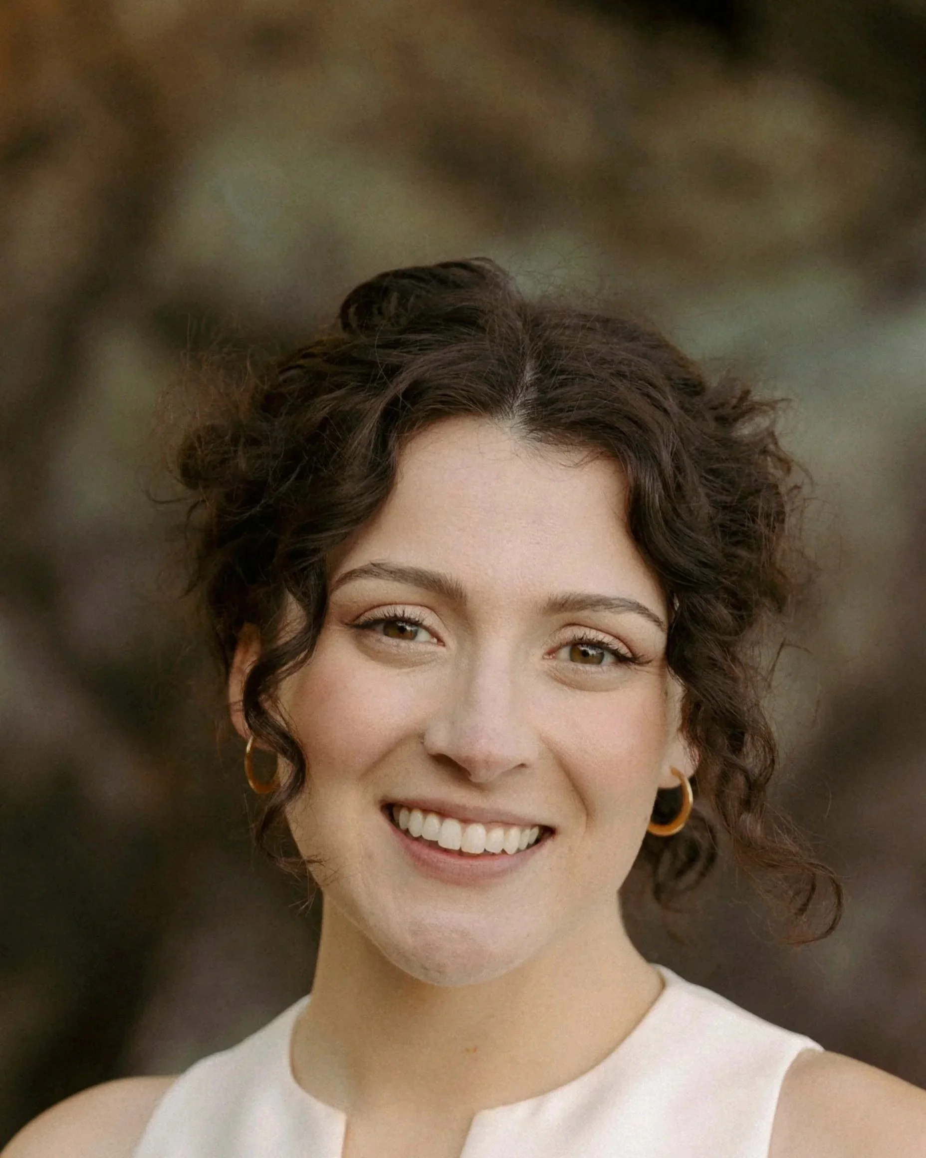 Close-up of a young woman with short curly brown hair, wearing gold hoop earrings, smiling, and dressed in a sleeveless white top.