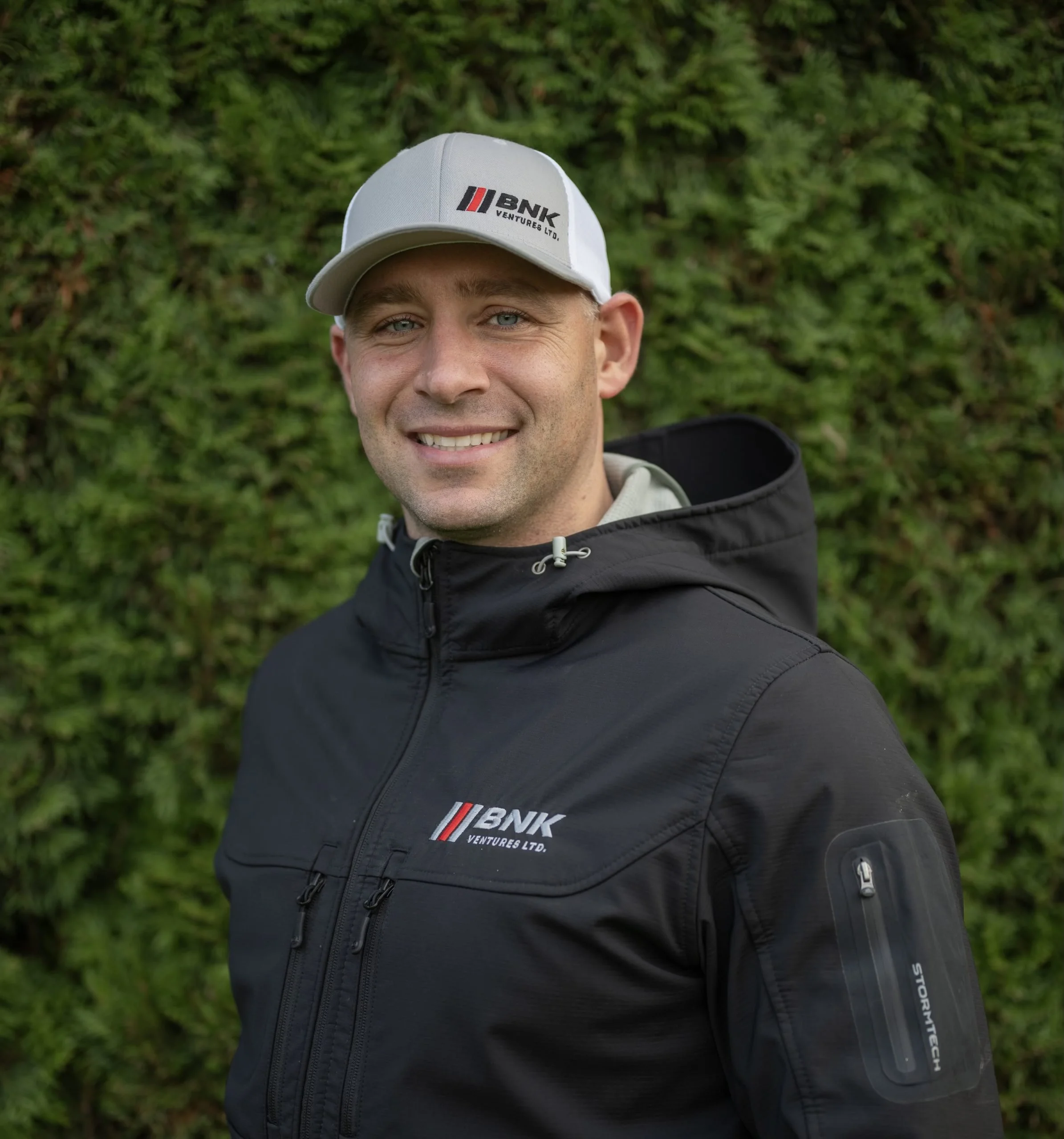 A smiling man in outdoor clothing, wearing a white cap and a black jacket with company logos, standing in front of a green hedge.