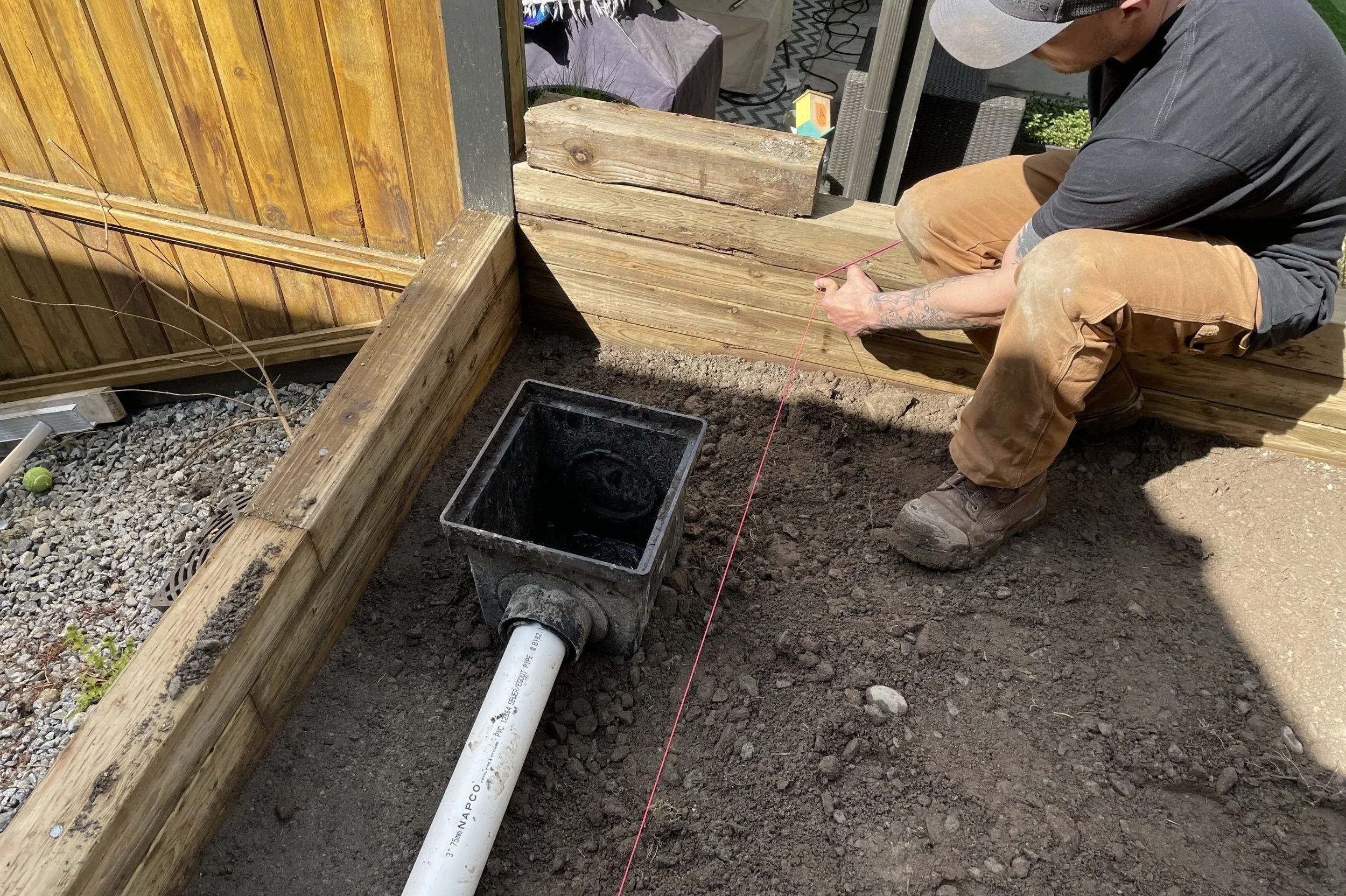 A person is working on a garden project, installing a drainage system with a pipe in a prepared soil area, surrounded by a wooden border.