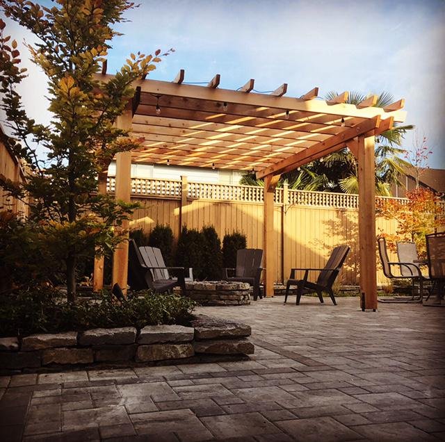 Backyard seating area with a wooden pergola, several chairs, a stone firepit, plants, and a fence.