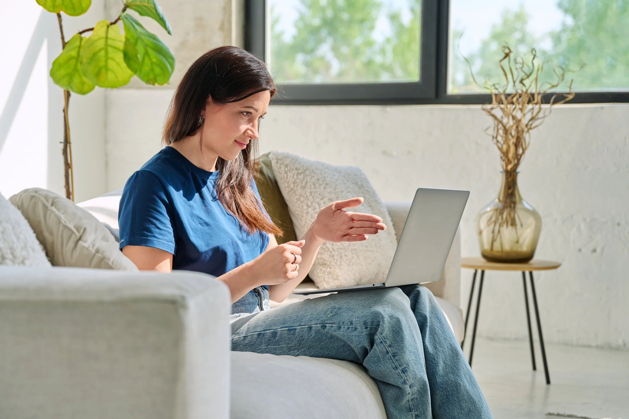 A woman sits on a sofa with a laptop on her lap, gesturing with her right hand, in a well-lit room with large windows, plants, and cozy pillows.
