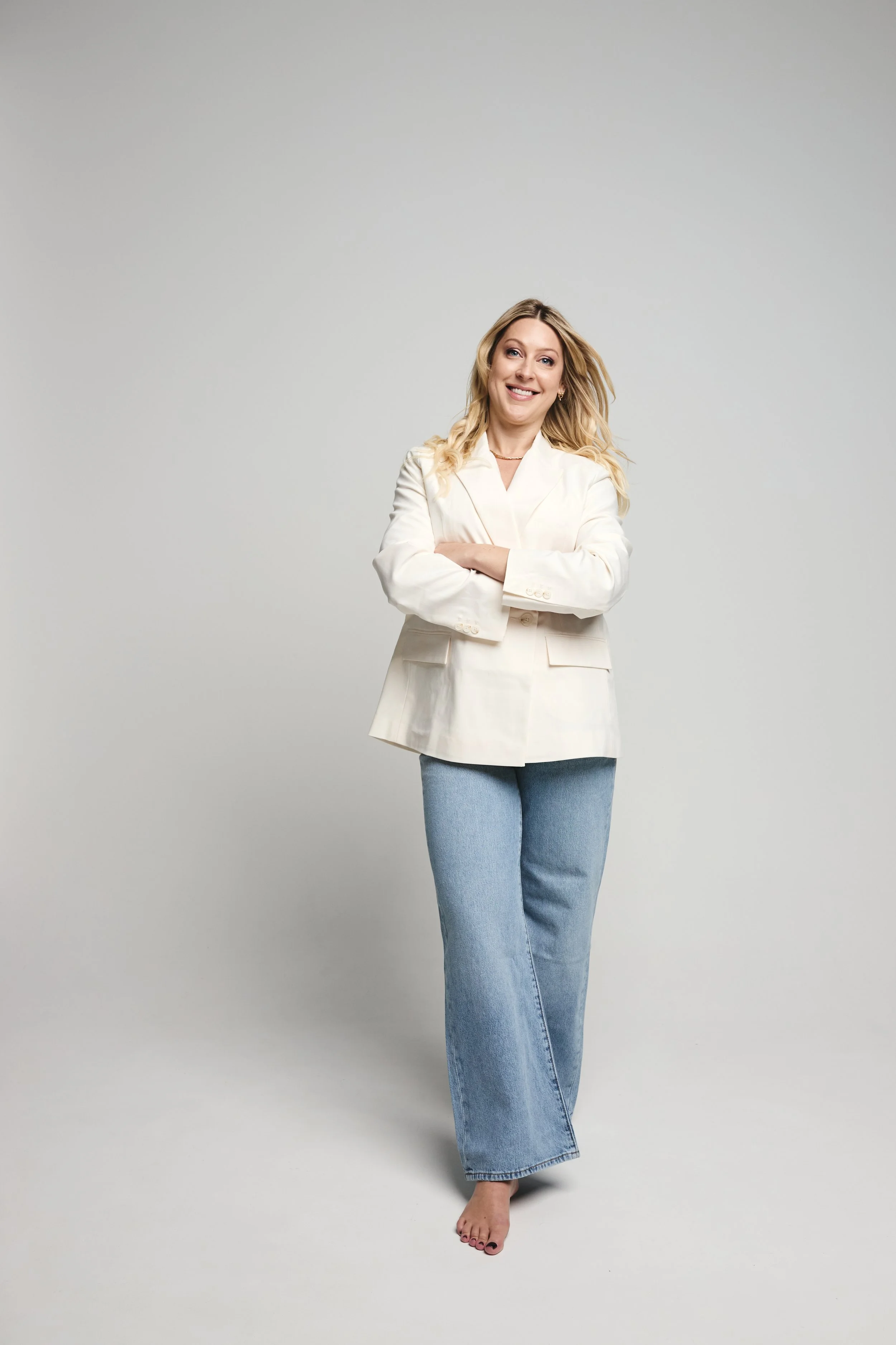 A woman with blonde hair wearing a white blazer and jeans, smiling and standing with her arms crossed on a plain light gray background.