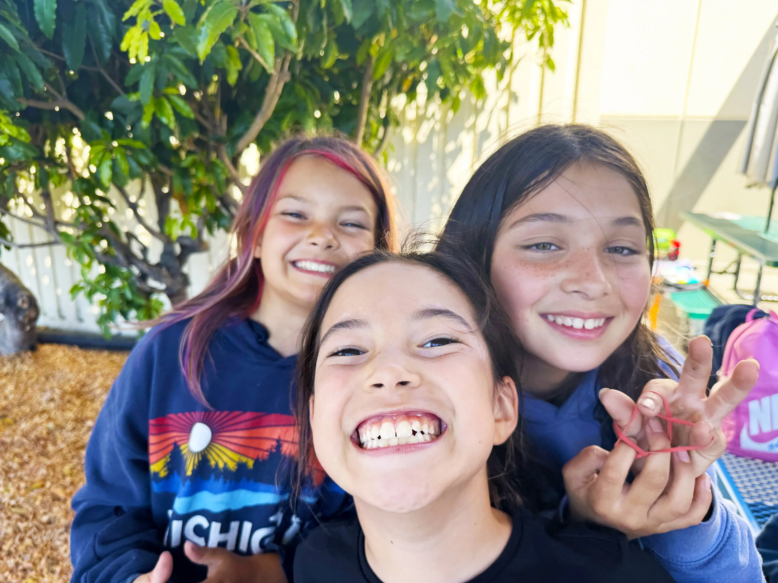 Three smiling girls taking a selfie outdoors with trees and playground equipment in the background.