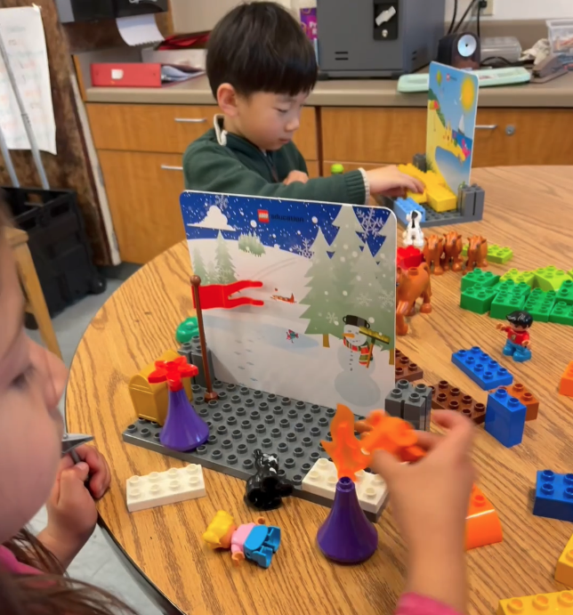 Children playing with LEGO bricks and toys on a wooden table, with a snowy winter scene backdrop and a cardboard cutout of trees and a snowman.