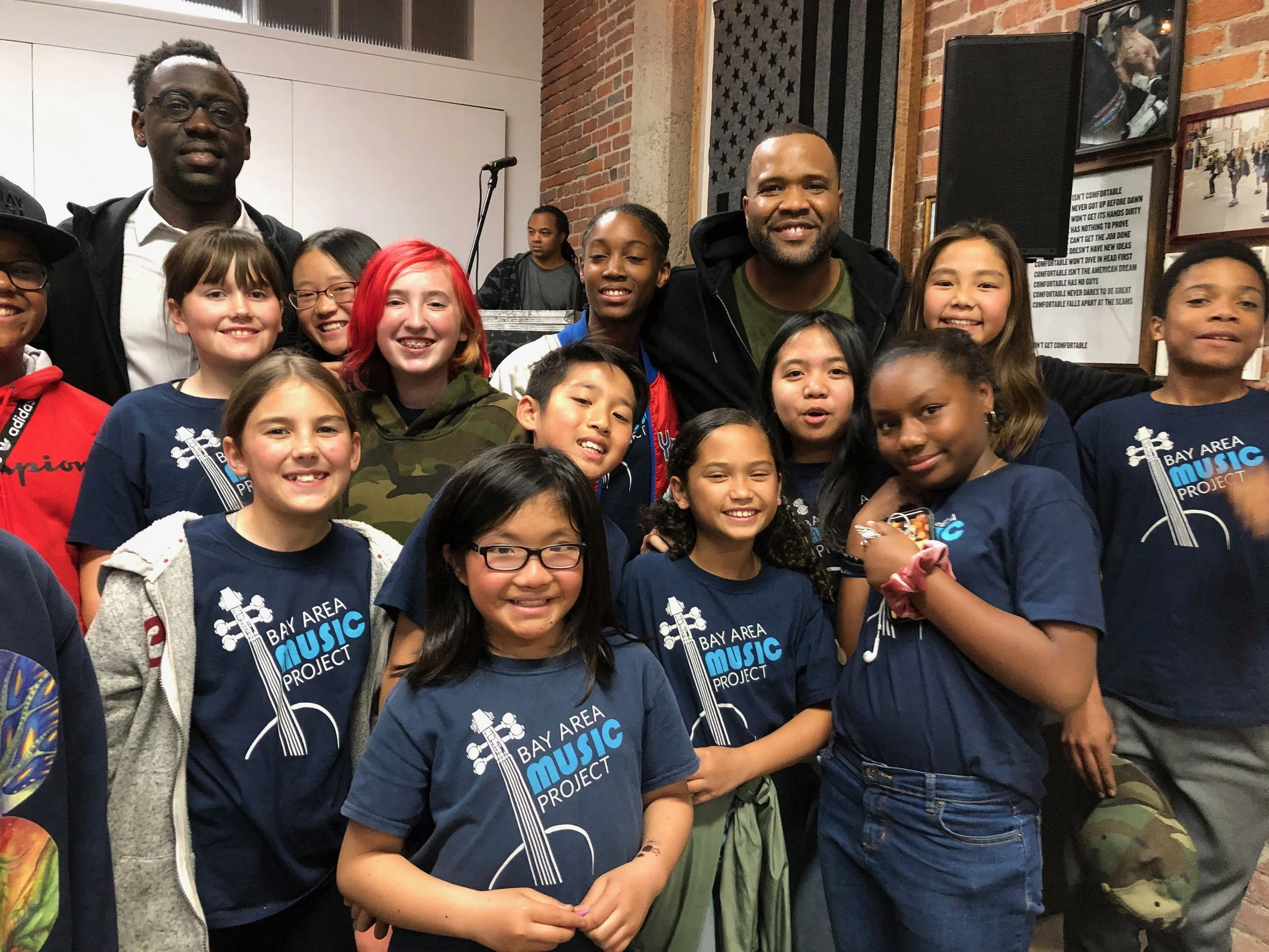 Group of children and two adults smiling and posing for a photo at the Bay Area Music Project event, with brick walls and framed pictures in the background.