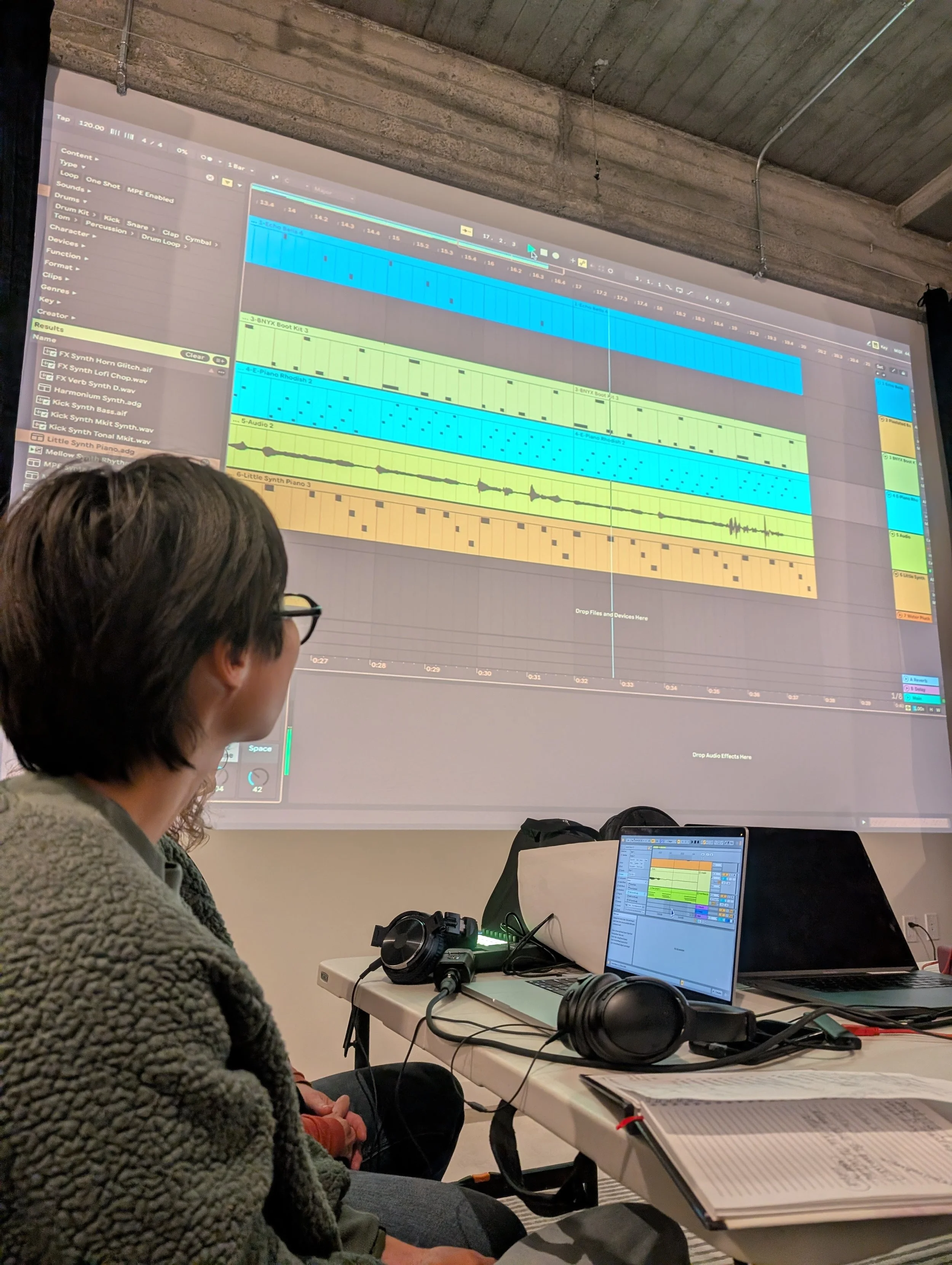 Person sitting in front of a large screen displaying digital music production software, with a laptop, headphones, and papers on the table.