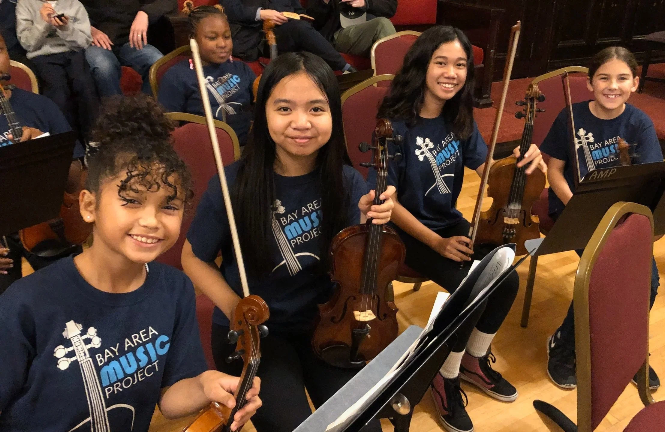 Group of young girls sitting with violins, smiling, in a music classroom or rehearsal space, with music stands and chairs around them, wearing matching navy blue shirts that say 'Bay Area Music Project.'