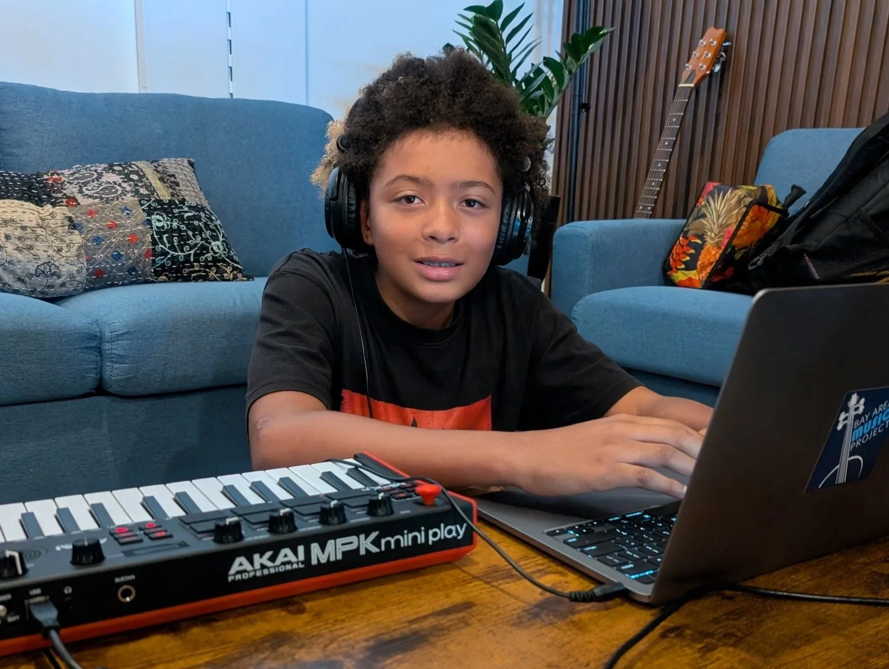 A young boy with curly hair wearing headphones is sitting at a wooden table, using a laptop, with a small MIDI keyboard/controller in front of him. There is a blue couch with decorative pillows and a guitar in the background.
