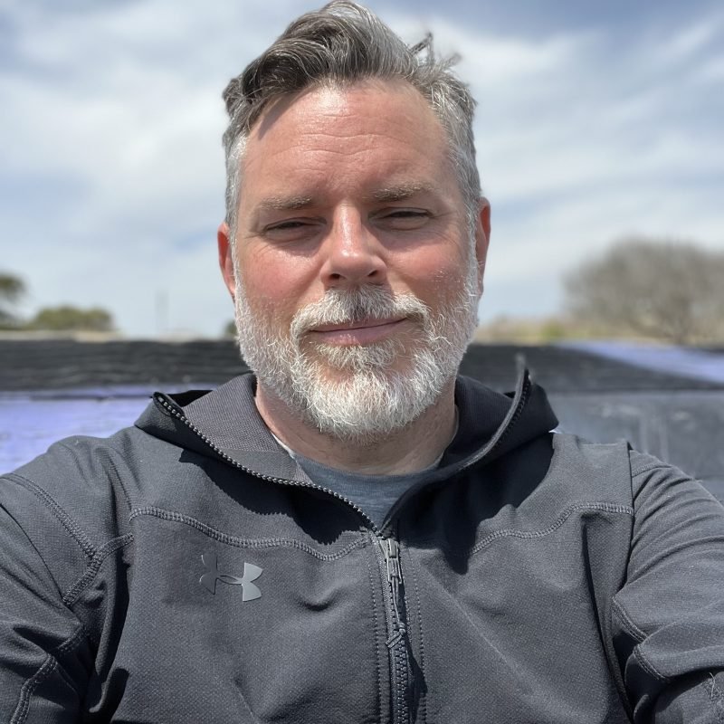 A man with gray hair and a beard takes a selfie outdoors on a cloudy day, wearing a black Under Armour jacket.