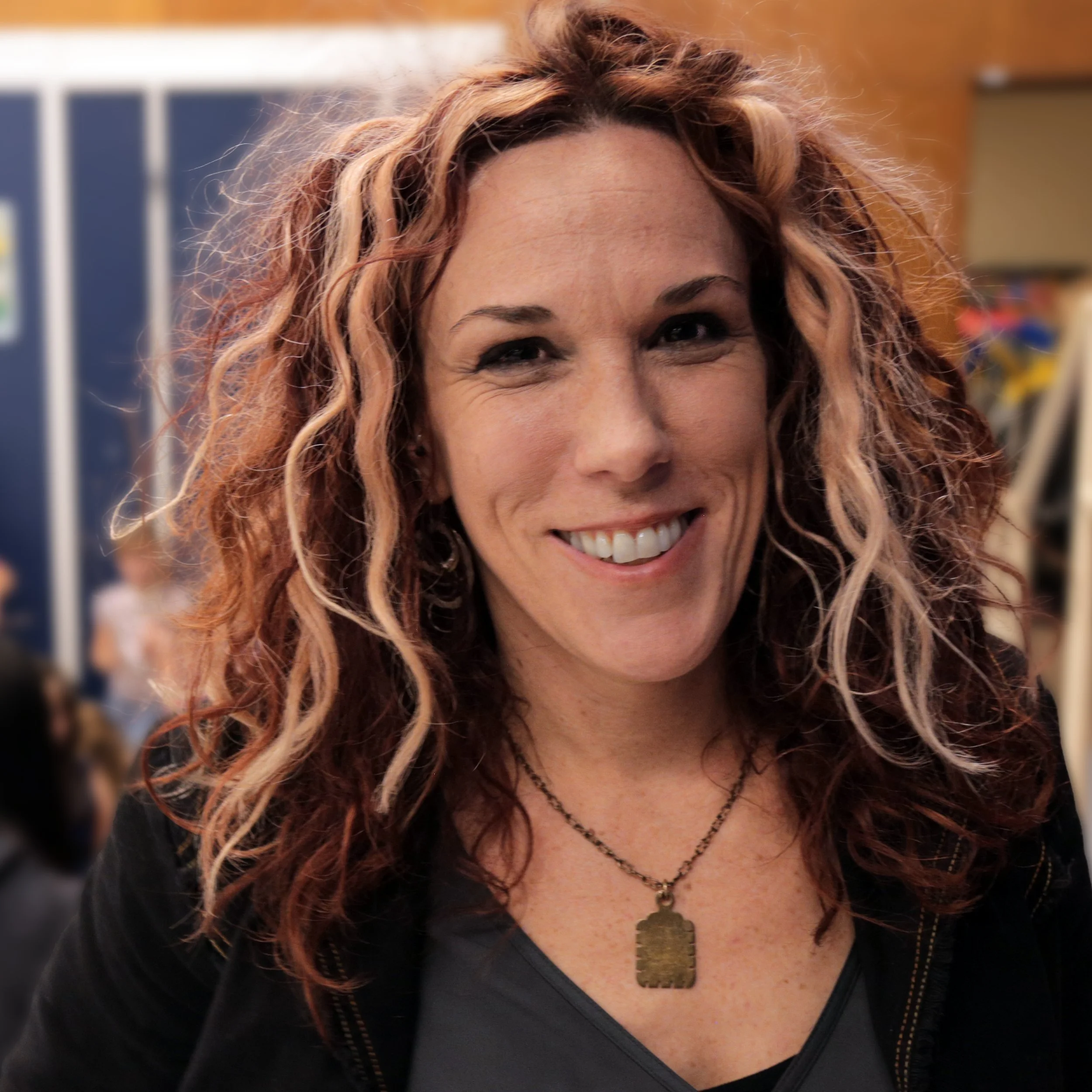 Close-up of Founder and Executive Director, Lorrie Murray with curly red hair, wearing a black jacket and a necklace with a pendant, in an indoor setting with others in the background.