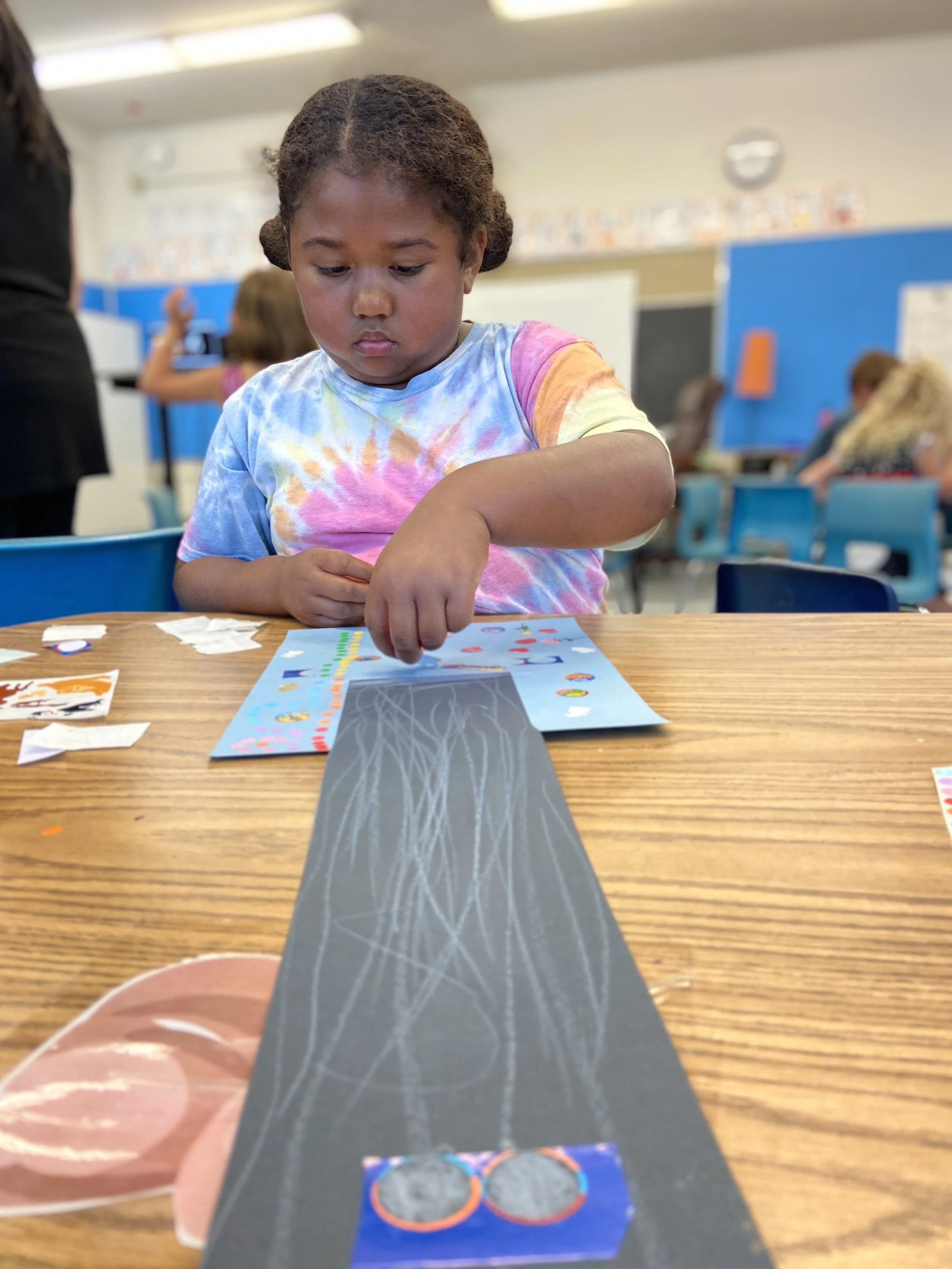 A young girl with braided hair wearing a colorful tie-dye shirt sits at a desk in a classroom, working on a craft project with stickers and paper.