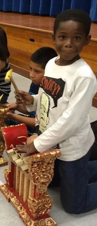 Two young boys participating in a cultural or religious event, with one boy holding a mallet and a red drum, and the other boy kneeling beside a decorated golden temple-like structure.