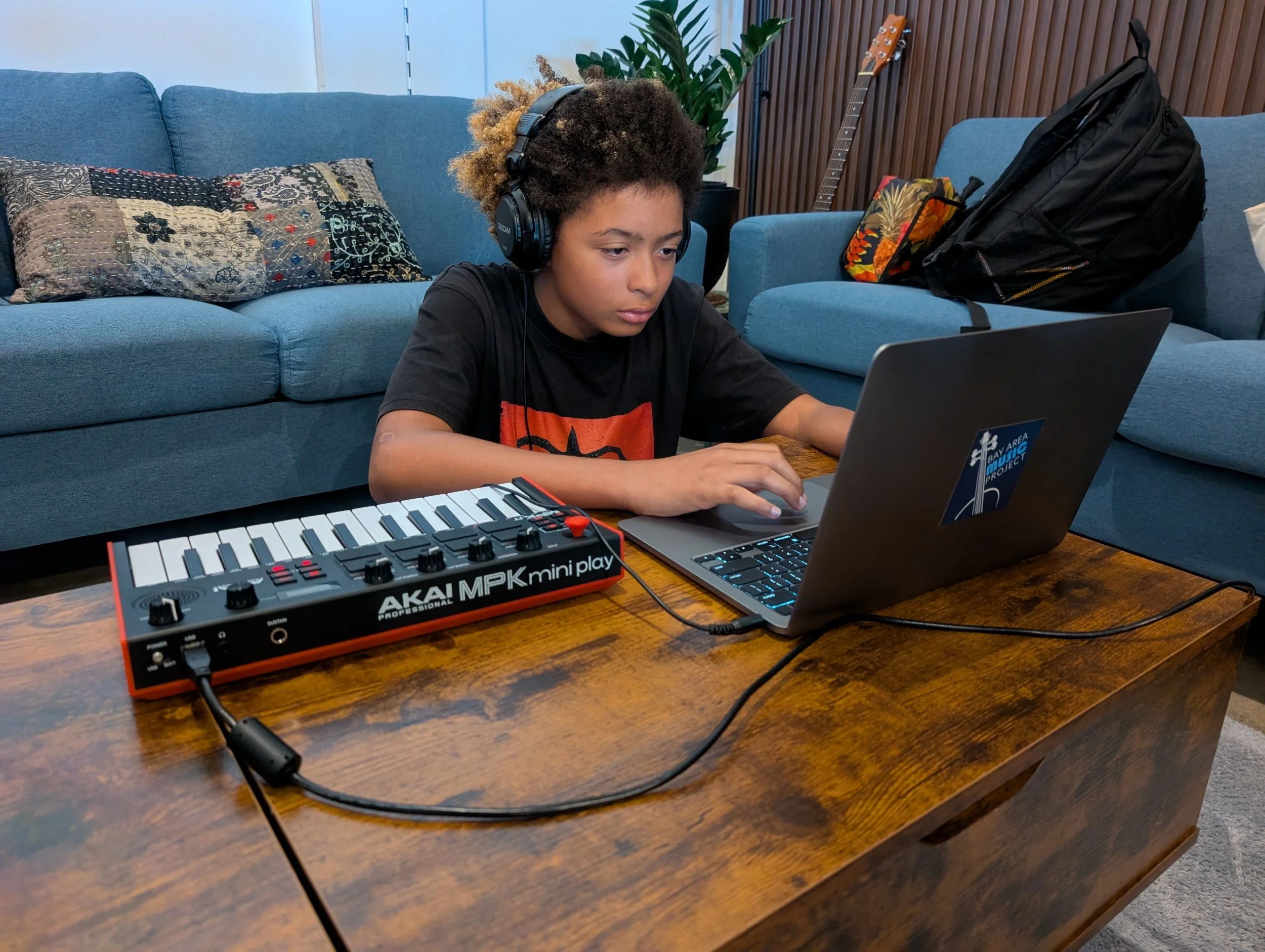 A young boy with headphones working on a laptop at a wooden table, with a music keyboard and a backpack in the background.