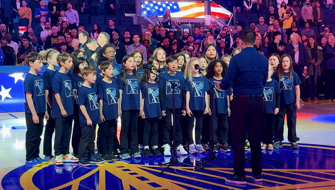 A children's choir performing on a basketball court with a conductor facing them, in front of an audience, with American flag-themed decorations in the background.
