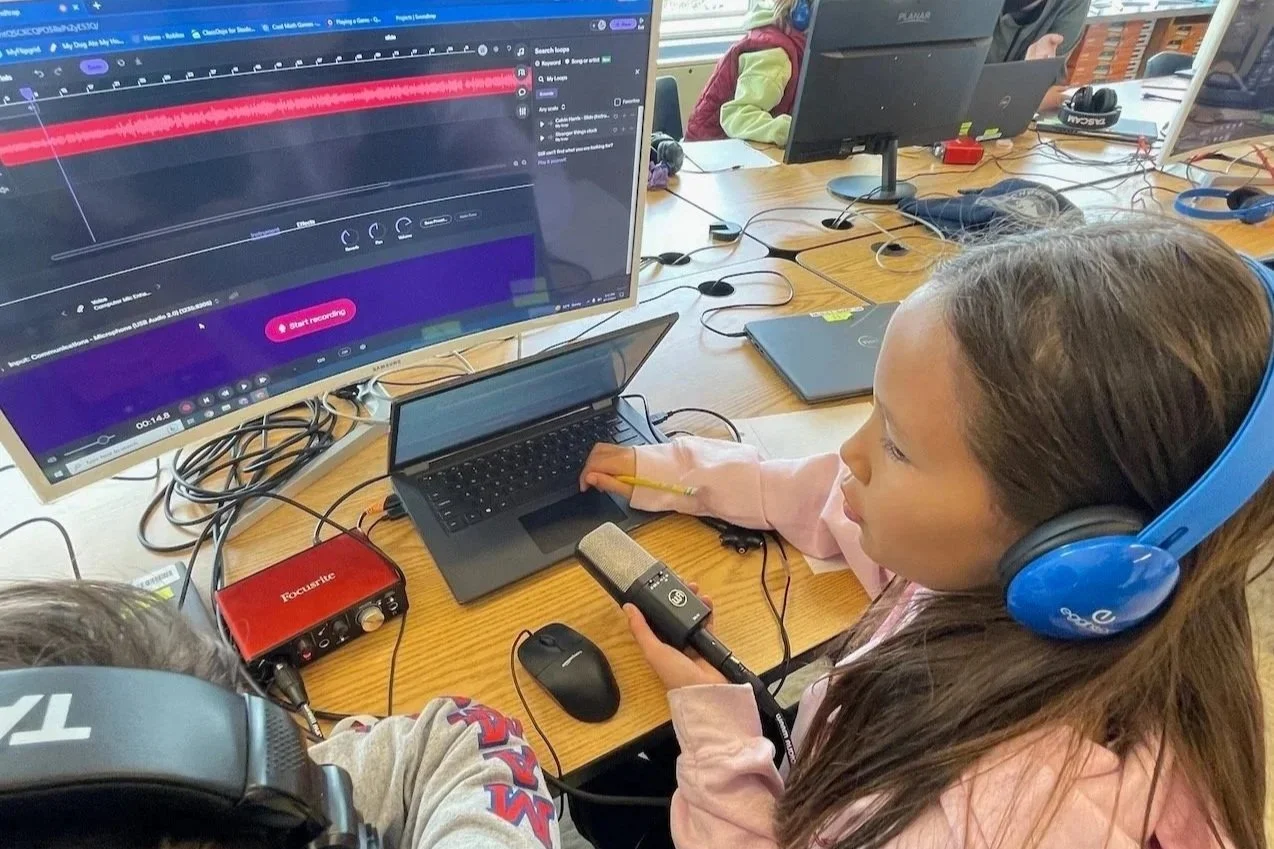 A young girl wearing blue headphones and holding a microphone sits at a desk with a laptop and desktop monitor, recording or editing audio or video as part of a classroom or computer lab setting.