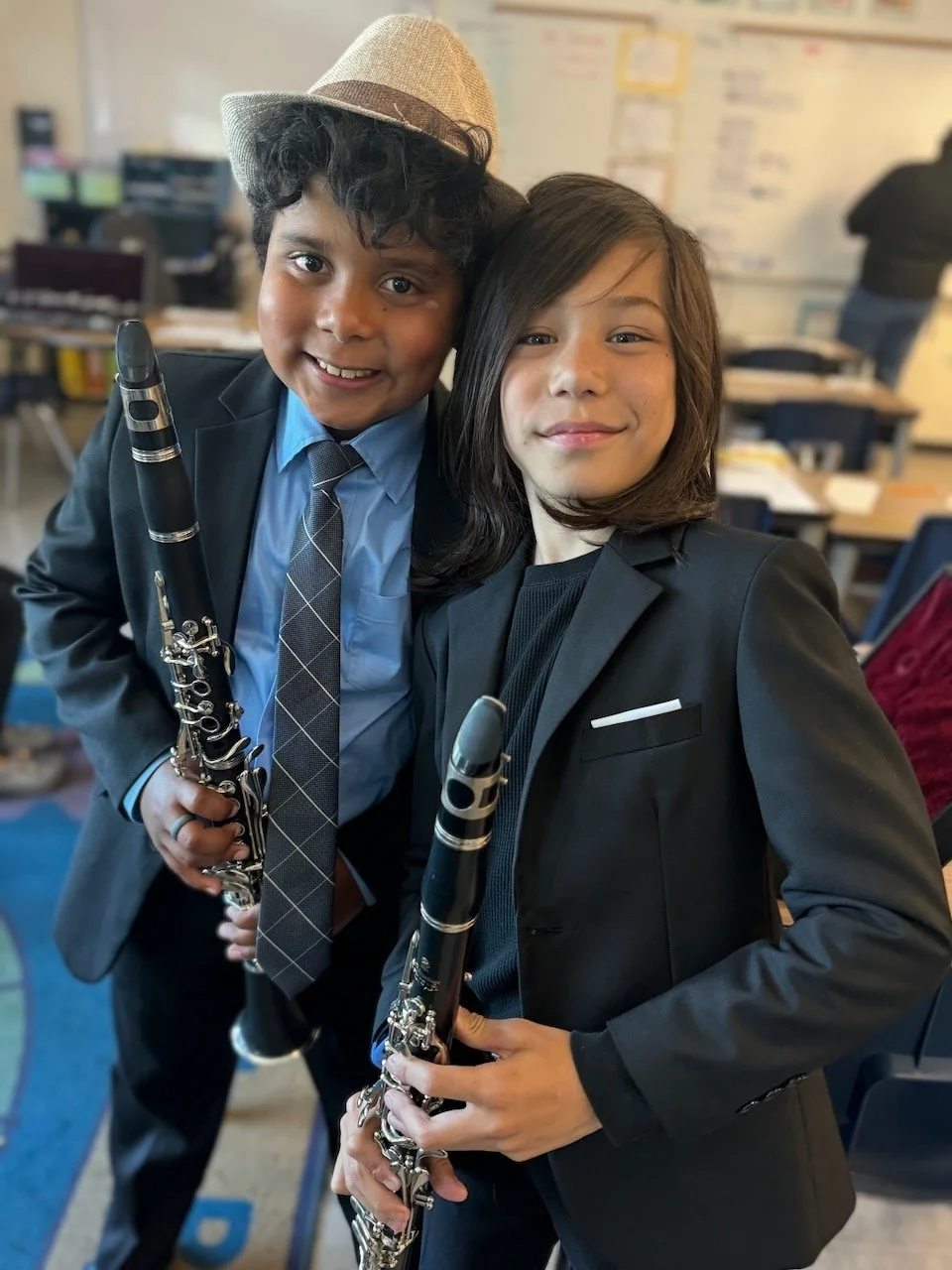 Two young boys in formal attire smiling and holding clarinets in a classroom.