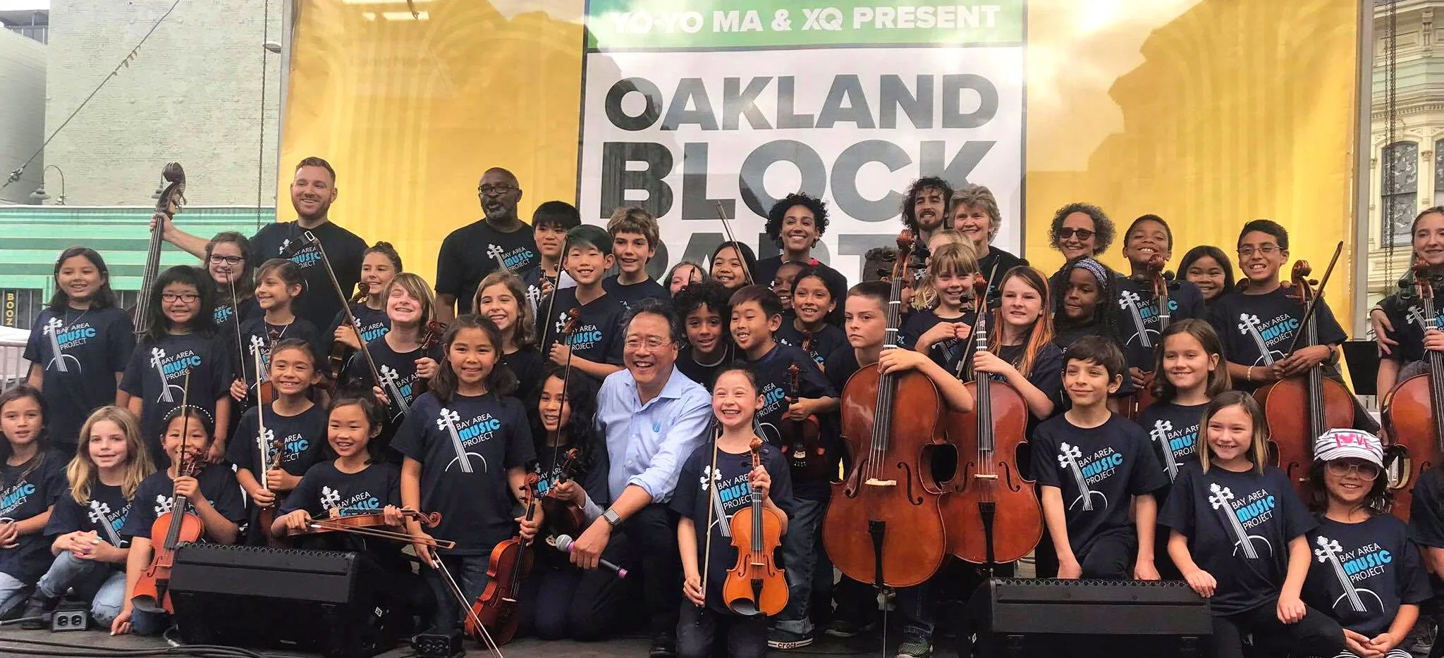 A large group of children and adults are on stage at an outdoor event called 'Oakland Block Party.' Most are holding musical instruments like violins and cellos, and are wearing matching dark blue shirts with 'Bay Area Music Project' printed on them. They are smiling and posing for a group photo.