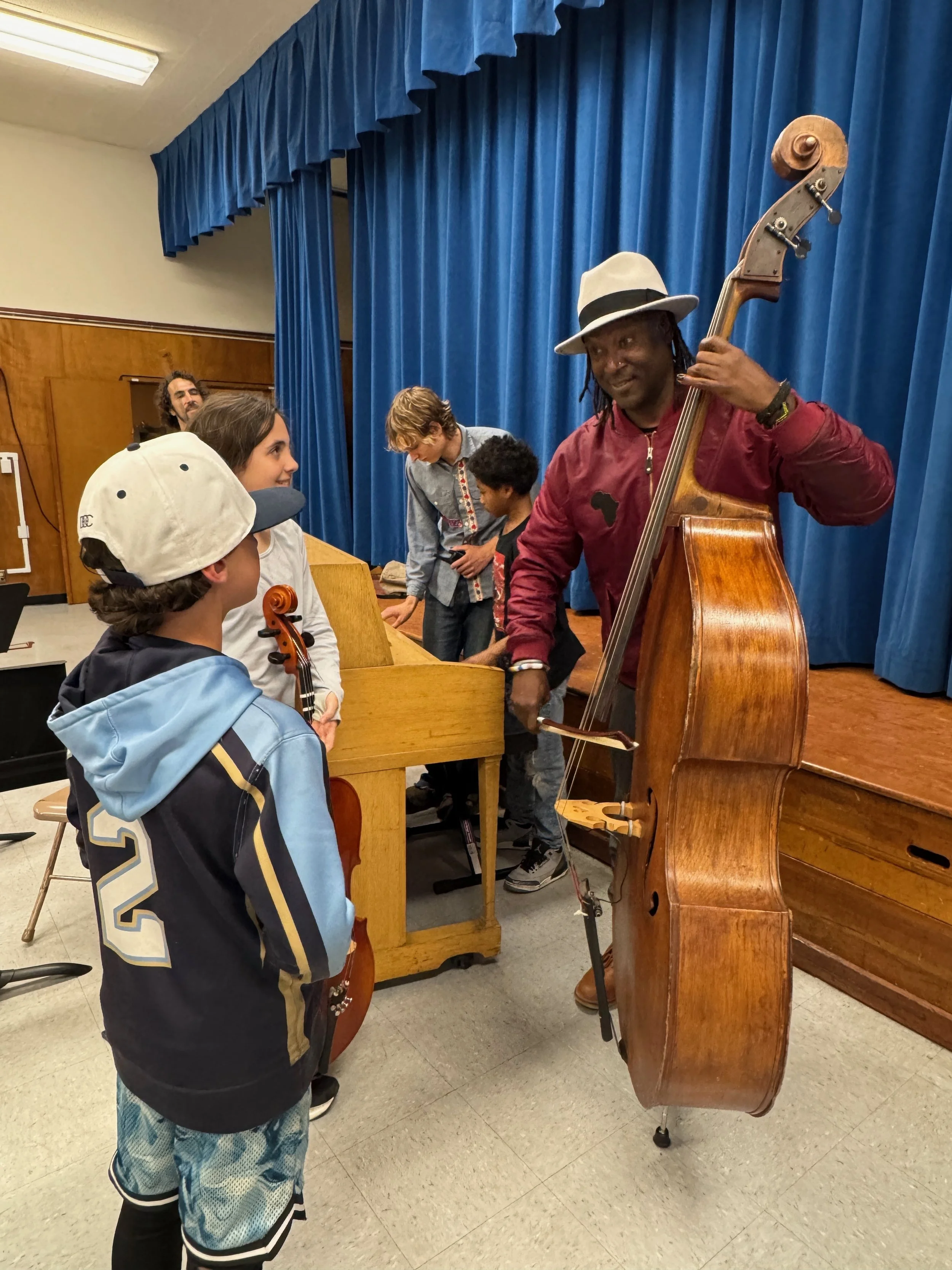 A man with a white fedora playing a double bass while talking to a young boy holding a violin. The scene takes place in a room with a blue curtain background, several children, and an upright piano.