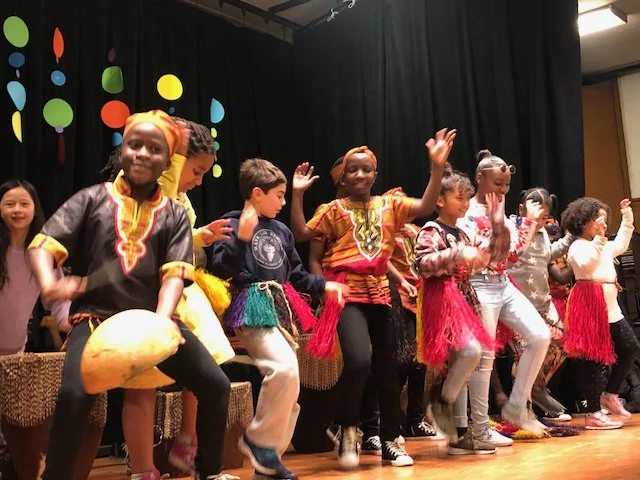 Children performing traditional dance on stage, dressed in colorful costumes, some with headwraps and skirts, with a black curtain and colorful polka dot decorations in the background.
