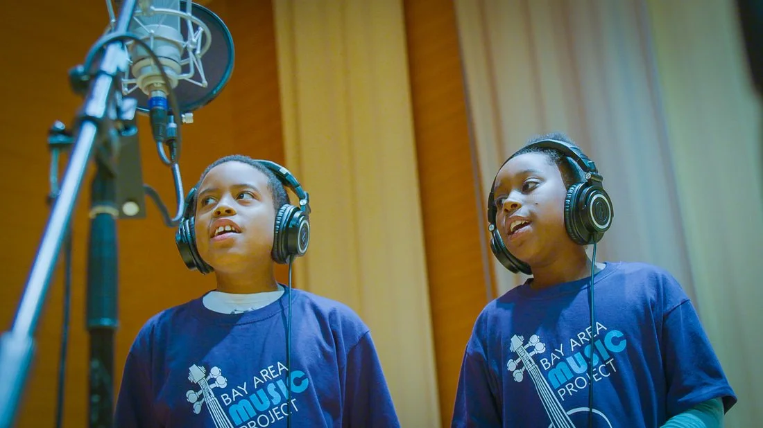 Two young children wearing blue T-shirts and headphones singing into a microphone in a recording studio.