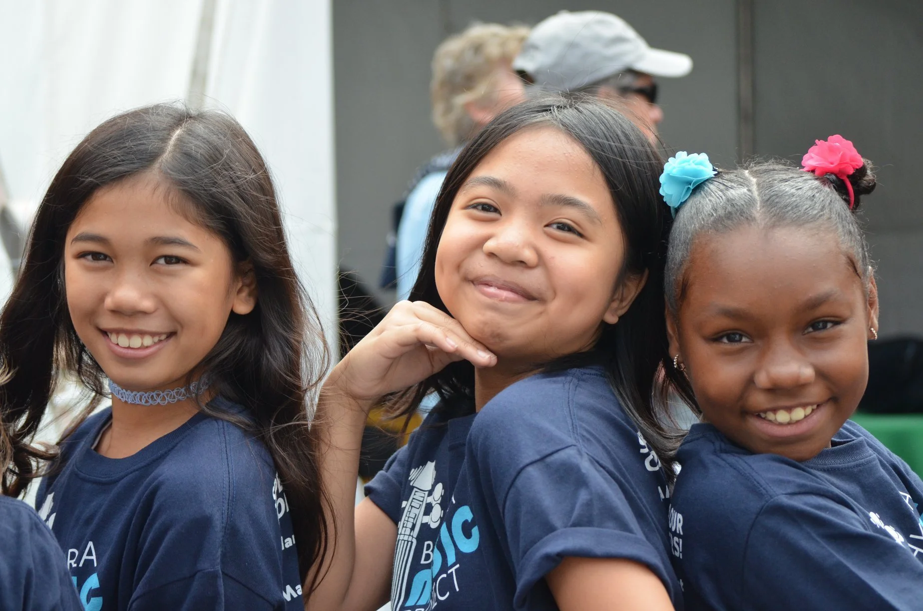Three smiling young girls with dark hair, wearing navy blue T-shirts, standing together outdoors, with two girls having colorful hair accessories and a woman with blonde hair and glasses in the background.