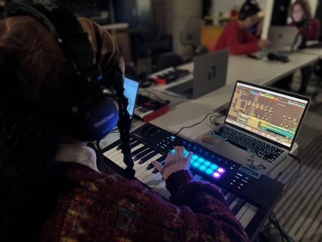 Person with braided hair and headphones playing a MIDI keyboard controller with a laptop displaying digital audio workstation software in a music production studio.