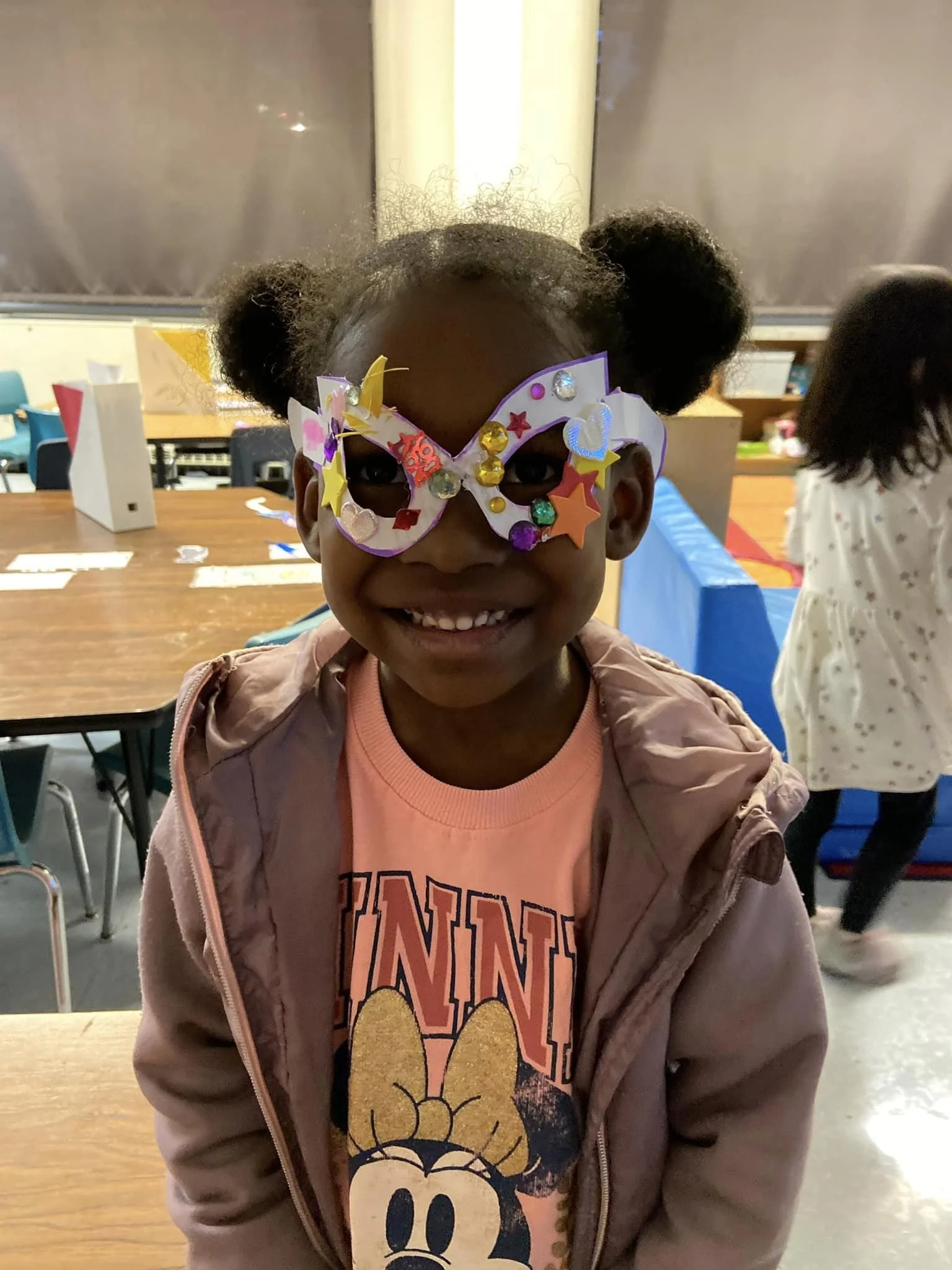 A young girl smiling while wearing colorful butterfly-shaped glasses decorated with glitter and stars, in a classroom or party setting.