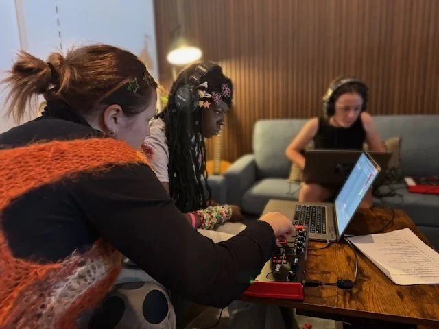 Three women are working with audio equipment and laptops at a wooden table in a cozy room. One woman is wearing headphones and adjusting sound mixer settings, while another women is typing on a laptop. The room has a gray sofa in the background.